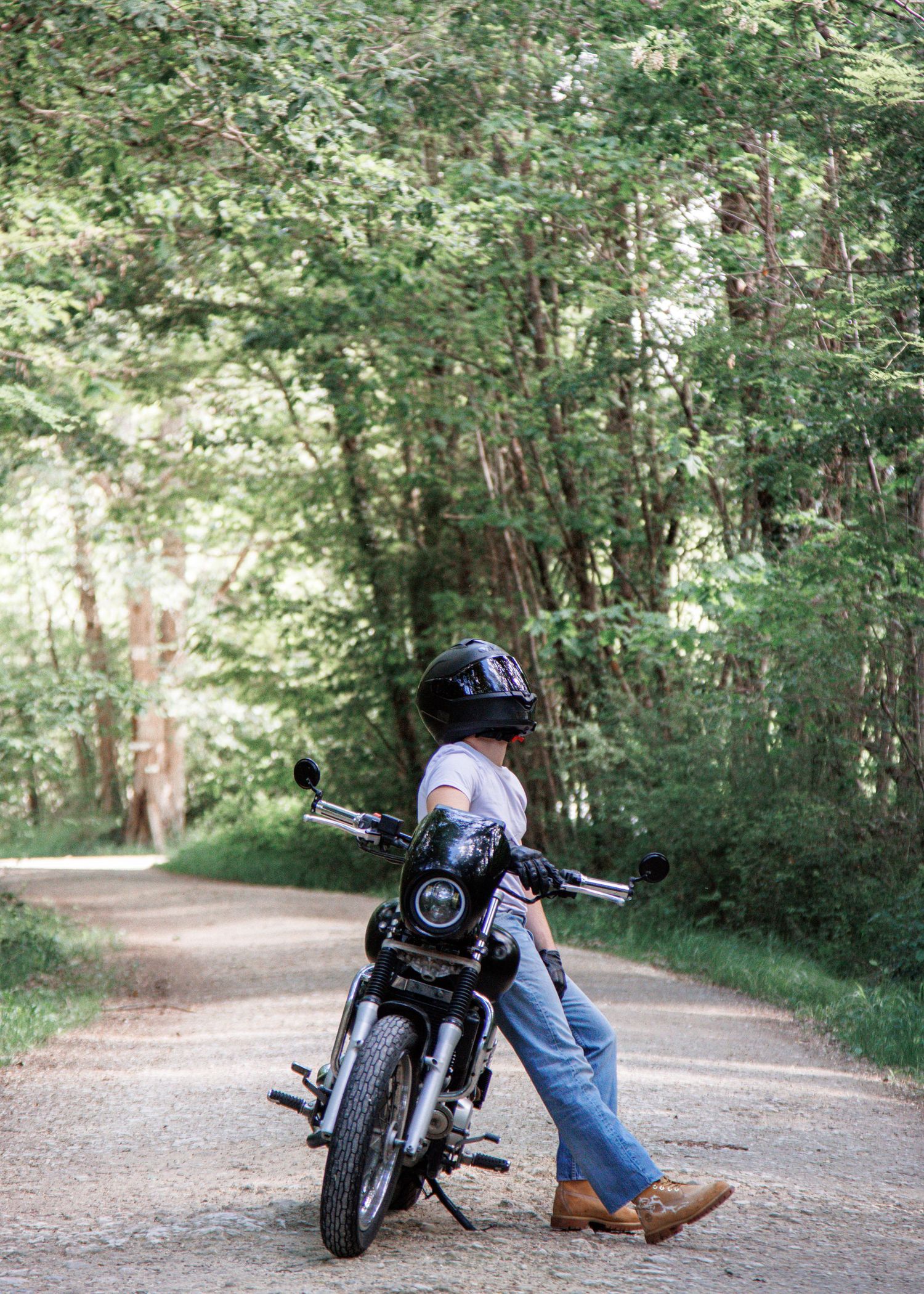 Person in helmet and jeans walking a black motorcycle down a tree-lined gravel road.