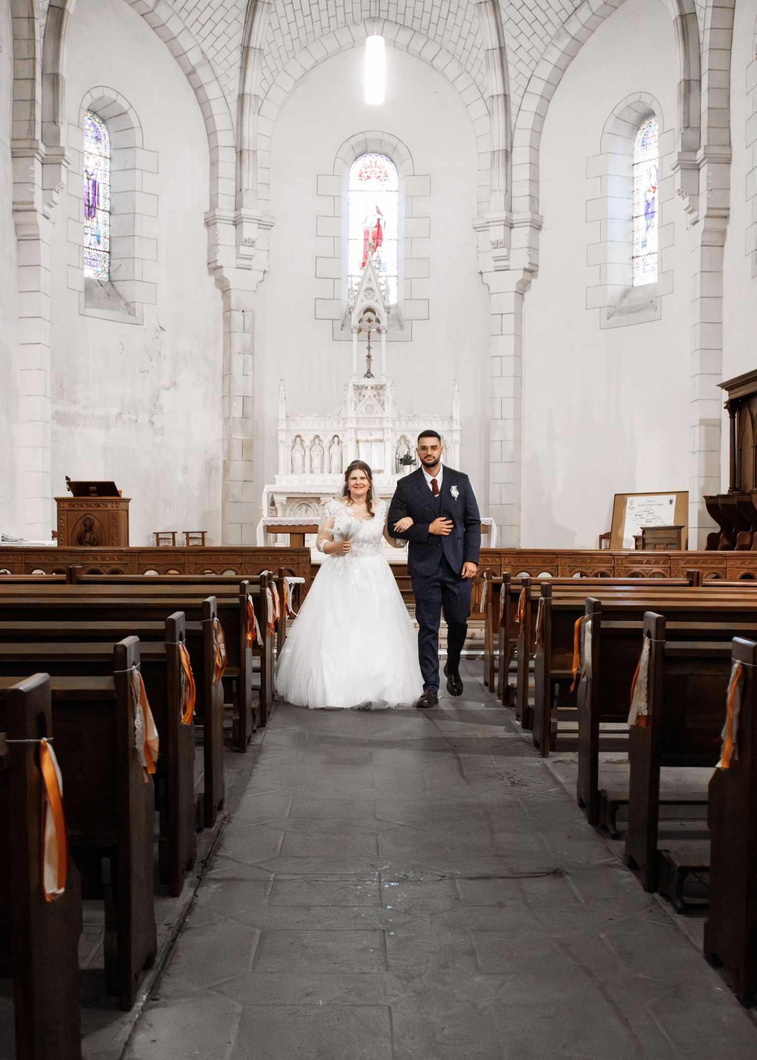 A bride and groom walk down the aisle of a beautiful white stone church with stained glass windows.