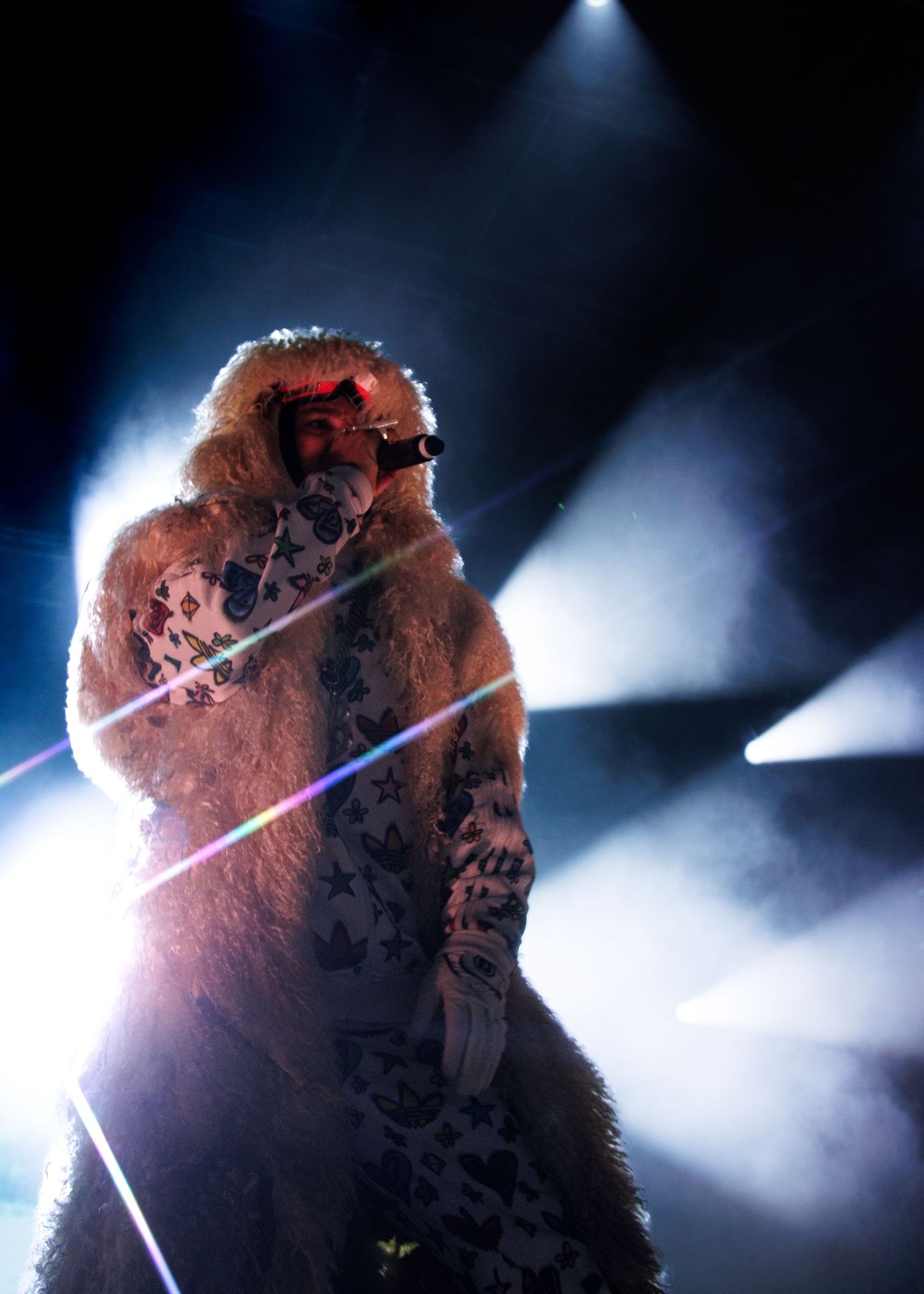A silhouetted figure in a fur coat stands dramatically backlit with beams of light streaming through smoke on a dark stage.