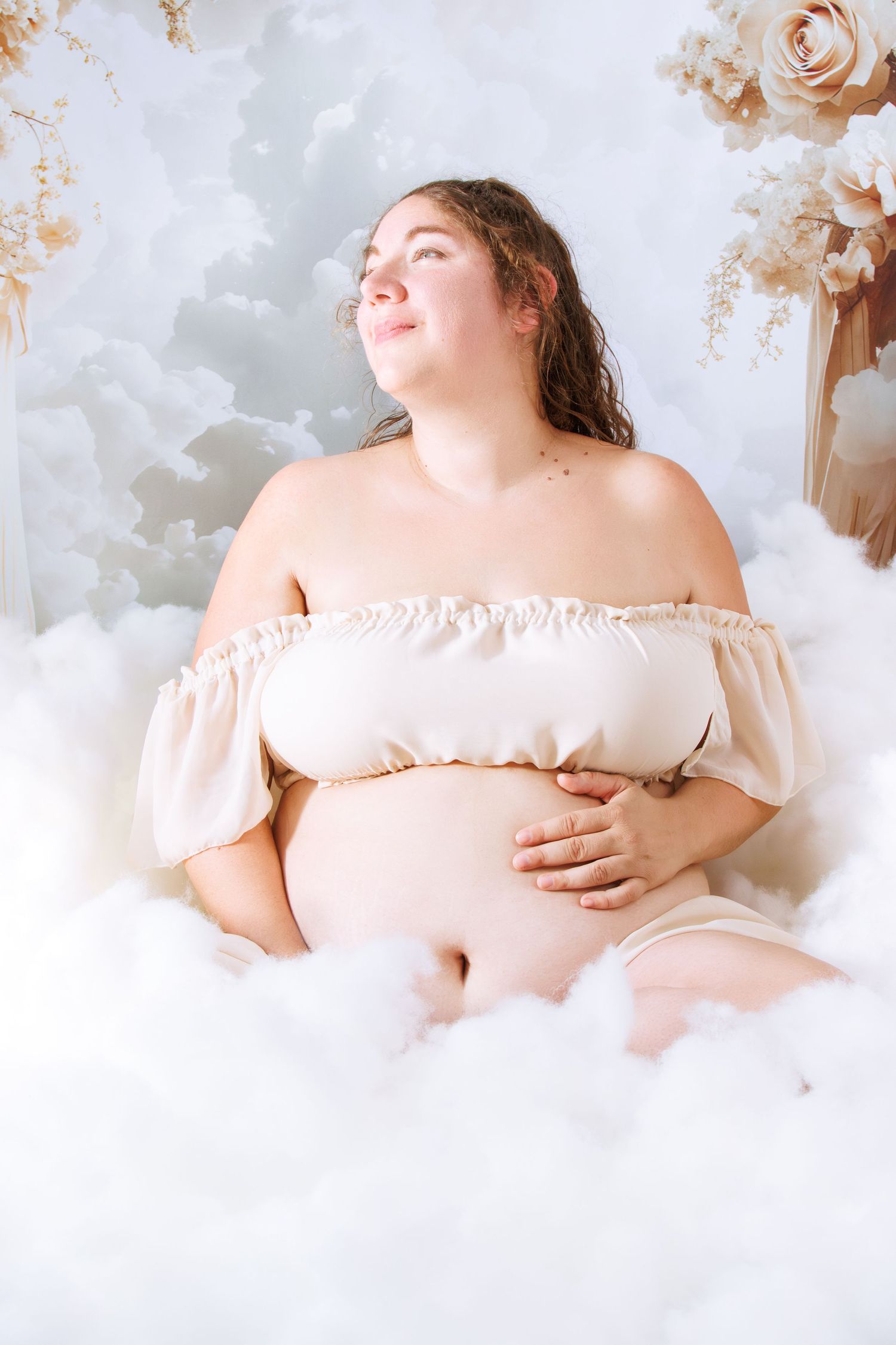 Romantic maternity photo with off-shoulder dress against cloudy white backdrop.
