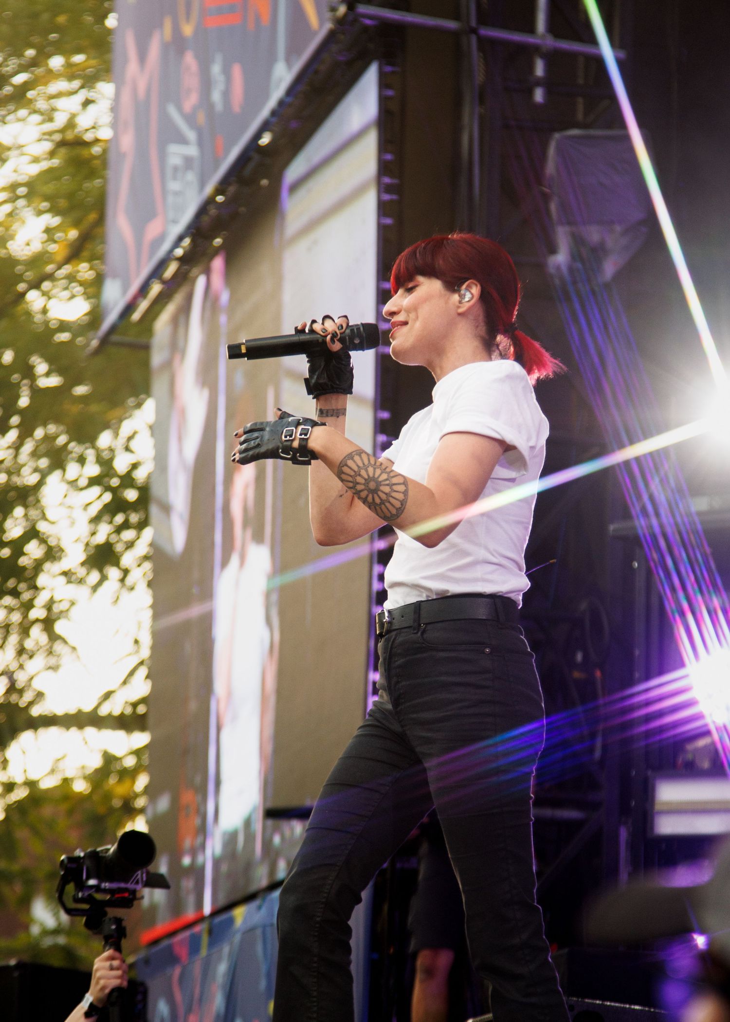 Red-haired female singer performs on stage with microphone, tattoos visible, wearing white tee and black jeans, colorful lasers behind.