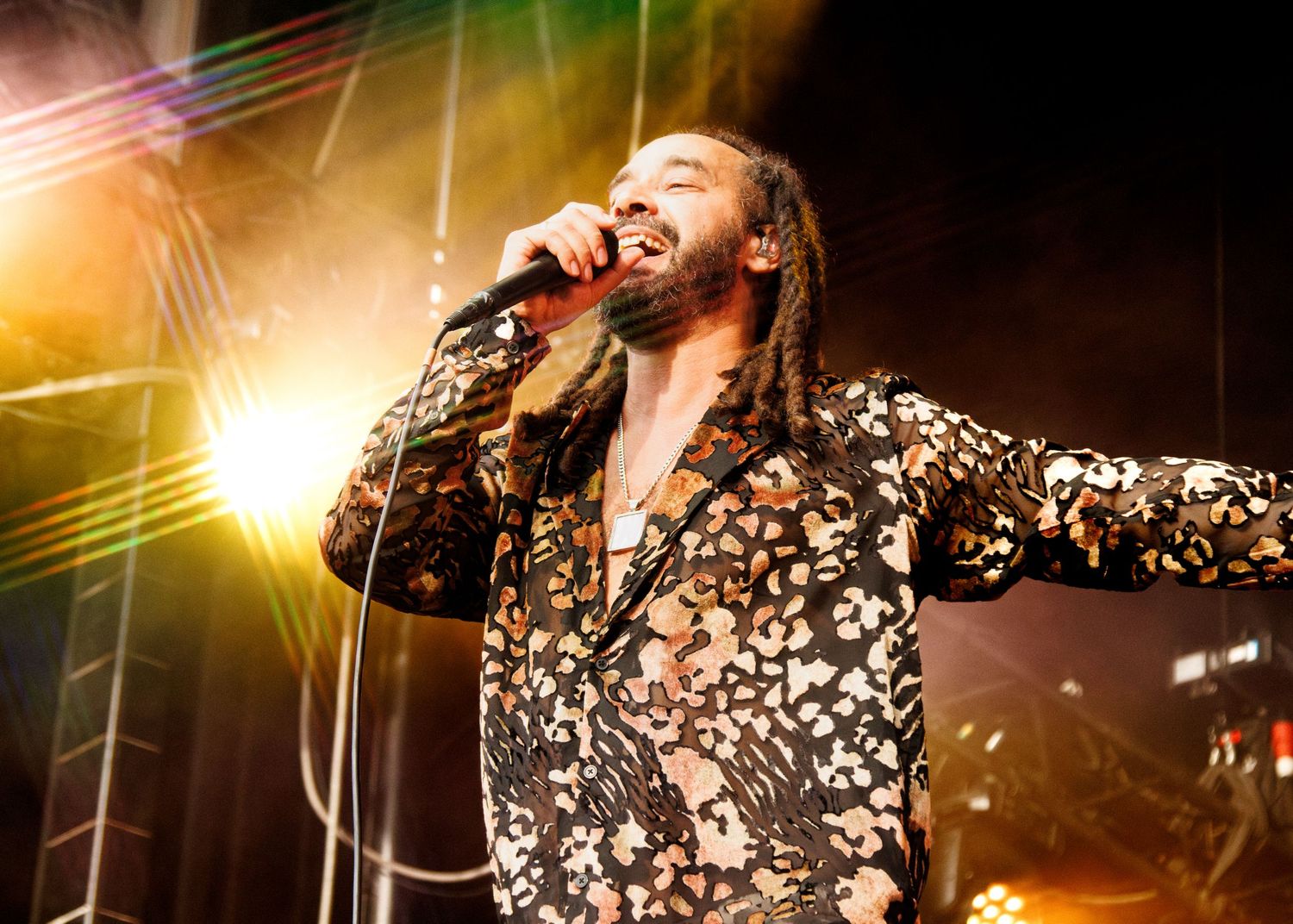 Reggae singer performing on stage with dreadlocks, floral jacket, and microphone under colorful stage lights.