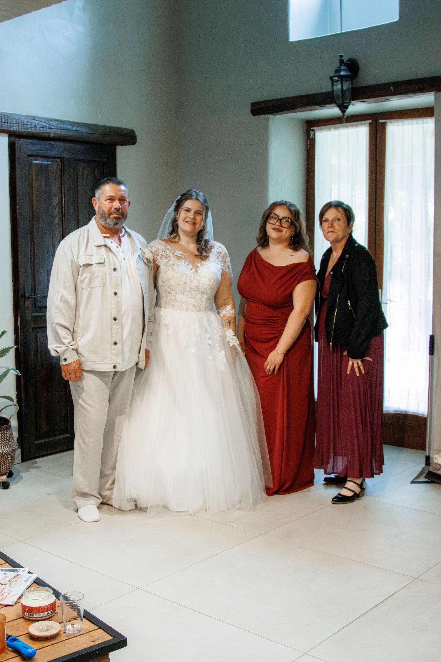 Bride in white lace gown poses with family in elegant venue, flanked by father and two women in red and black.