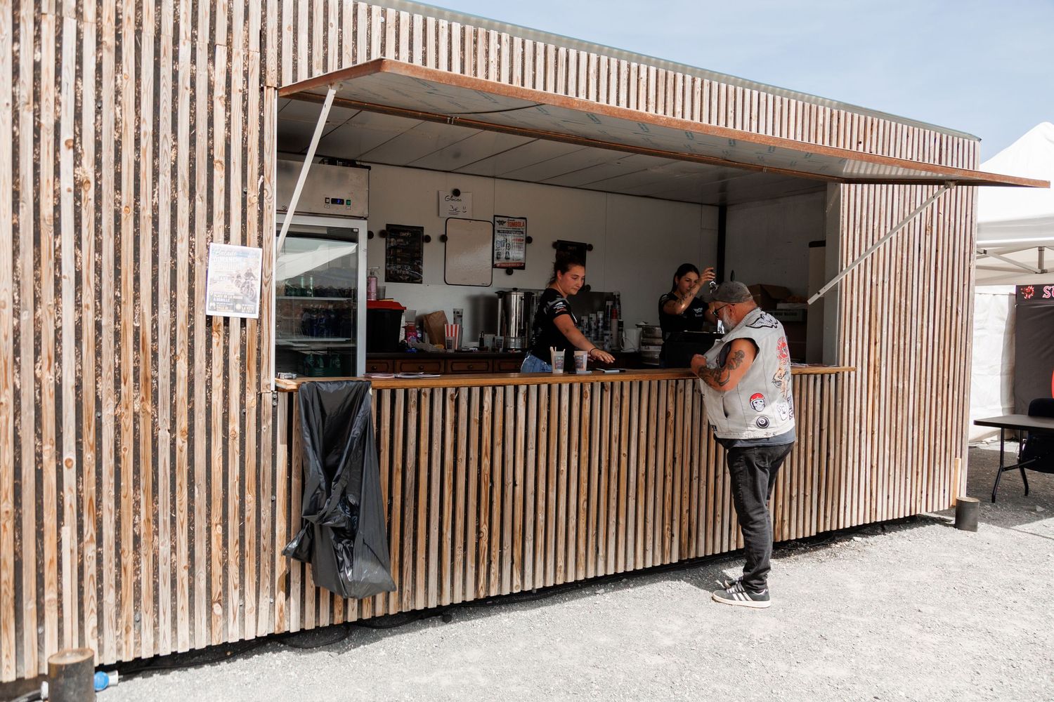 Outdoor food stall with wooden slat facade, two servers behind counter assisting a customer on a sunny day.