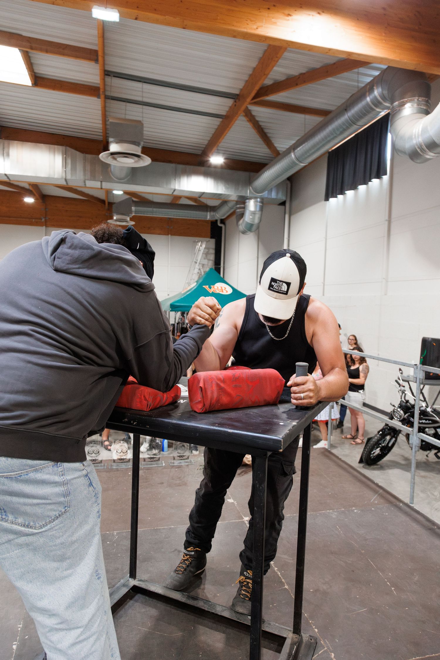 Two men arm wrestling on a padded table in an indoor venue, one wearing a North Face cap and black tank top.