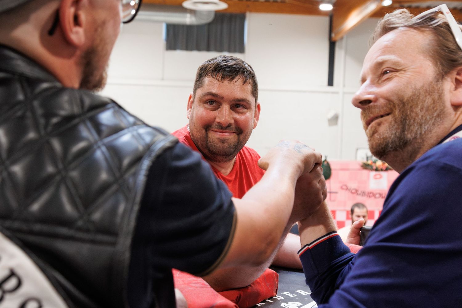 Three men smiling and arm wrestling at an indoor event, with one wearing a red shirt in the center.