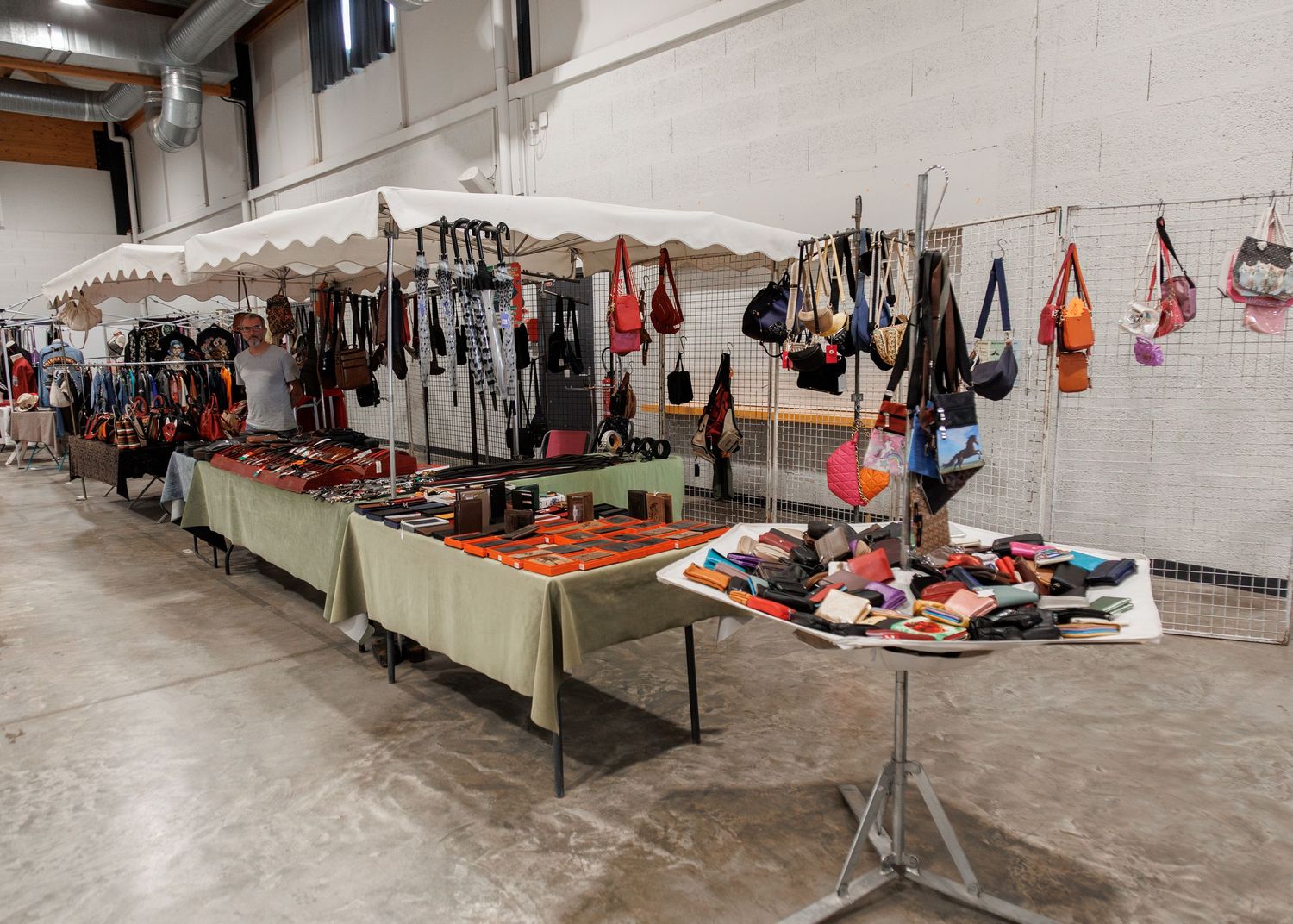 Indoor market stall displaying leather bags, purses, and accessories on tables and hanging racks in a warehouse venue.