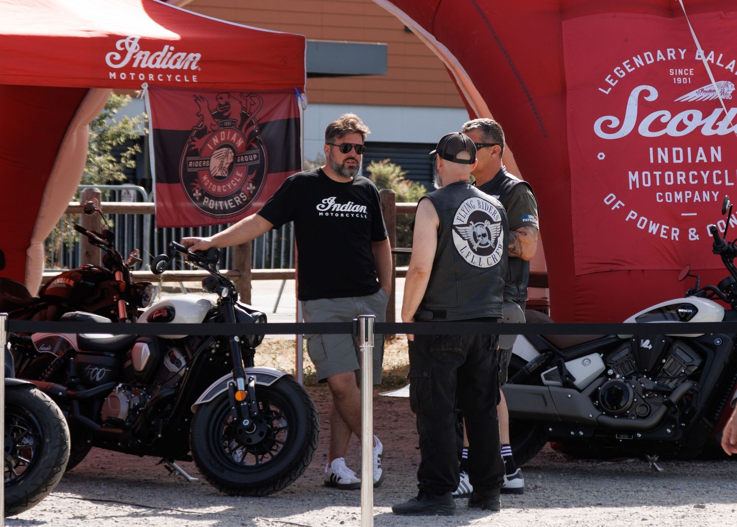 Two men discussing Indian Motorcycle models at an outdoor dealership event with Scout and Chief bikes on display.