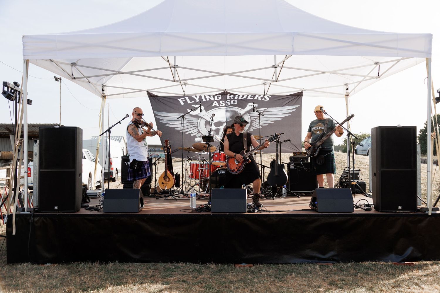 Live band performs on outdoor stage under white canopy tent with speakers and banner at sunny festival event.