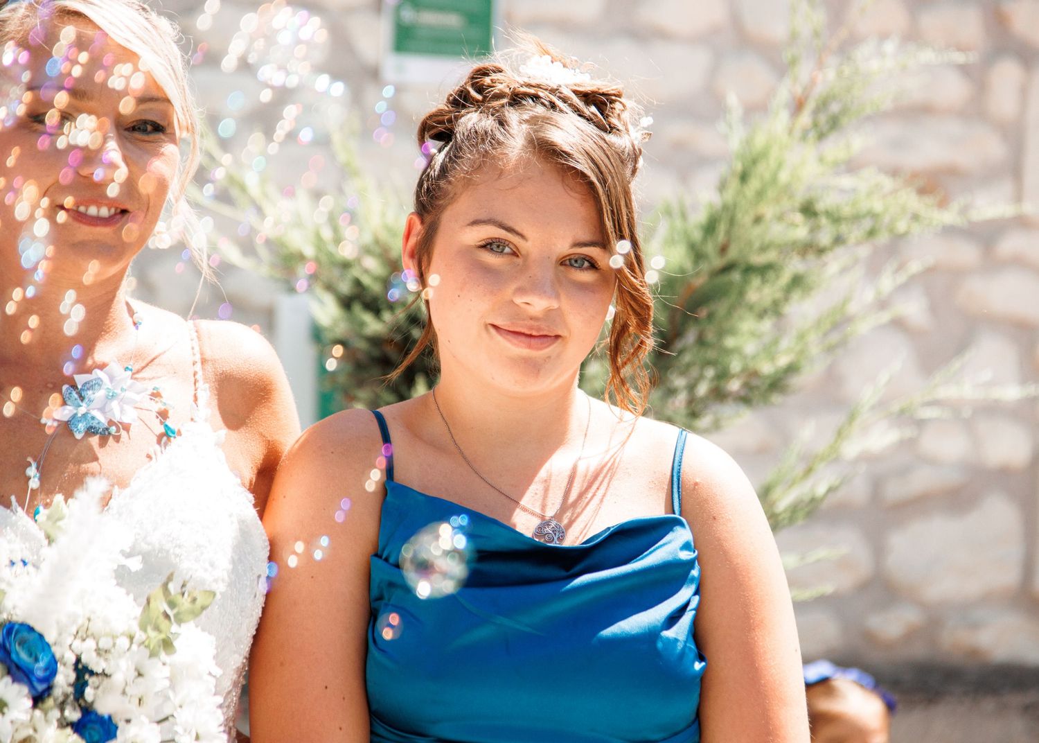 A guest in a blue dress smiles during an outdoor celebration with bubbles floating around.