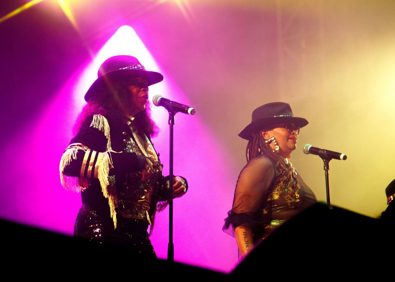 Concert performers sing onstage amid dramatic pink and yellow lighting effects during a live music performance.