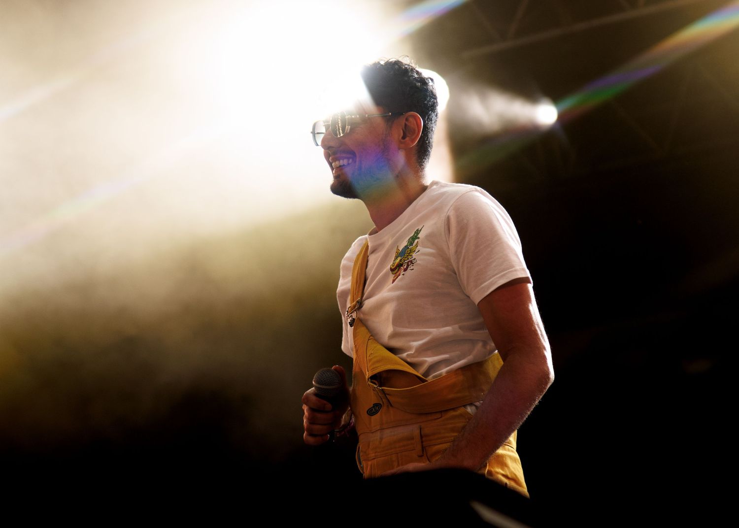 A performer on stage wearing a white t-shirt and brown vest stands in dramatic backlighting with light beams visible.