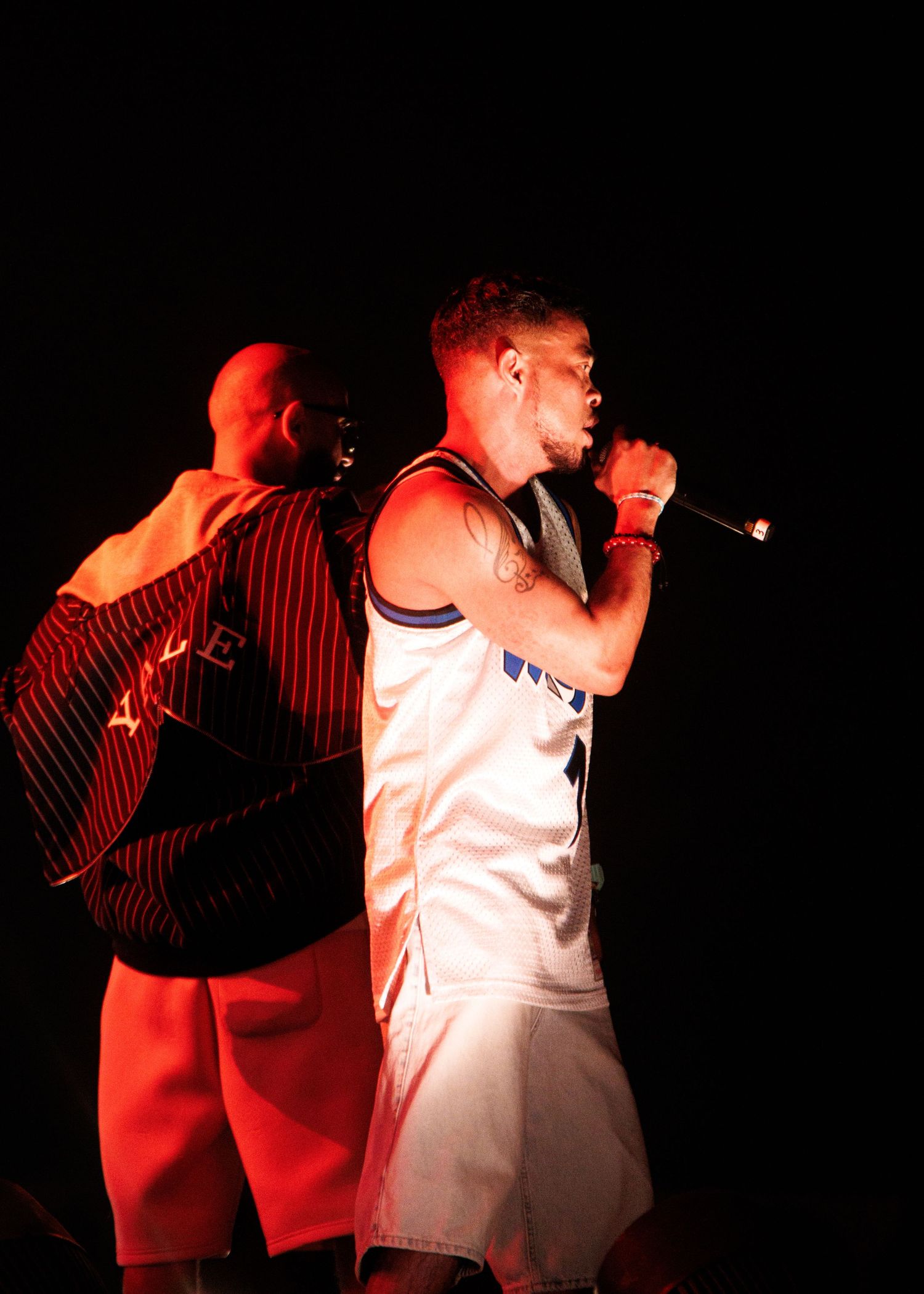 Two performers on a dark stage with one in a white basketball jersey holding a microphone during a live show.