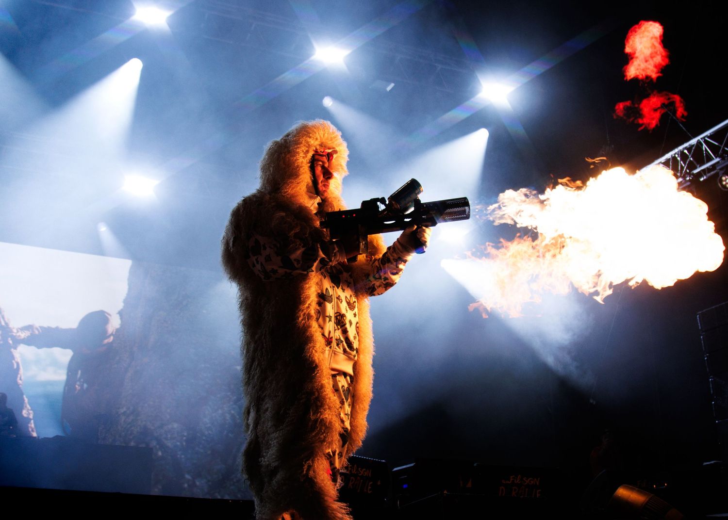 Metal band performer breathing fire on stage during a dramatic concert performance with blue lighting and smoke effects.