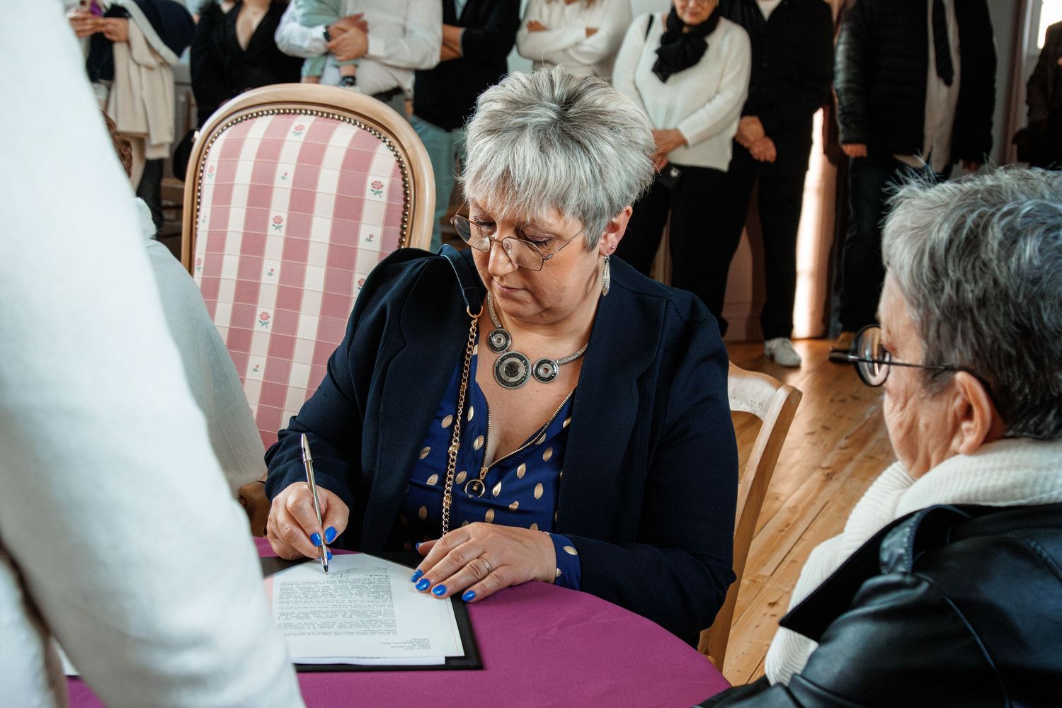 Elderly woman with short gray hair signing a document at a purple-draped table during a formal ceremony.