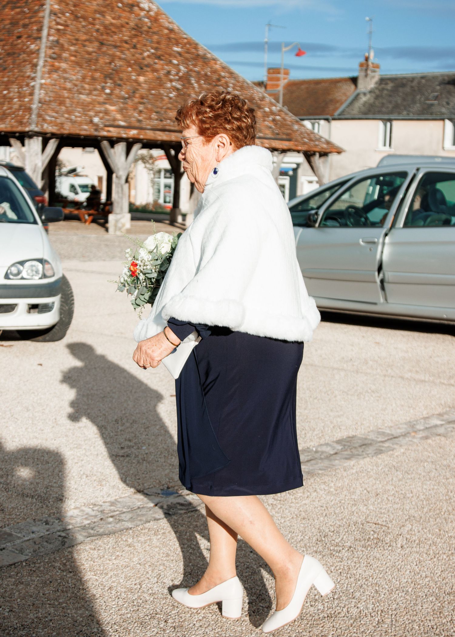 Elderly woman in white fur shawl, navy skirt, and white heels holding a bouquet, walking in a sunny outdoor setting.