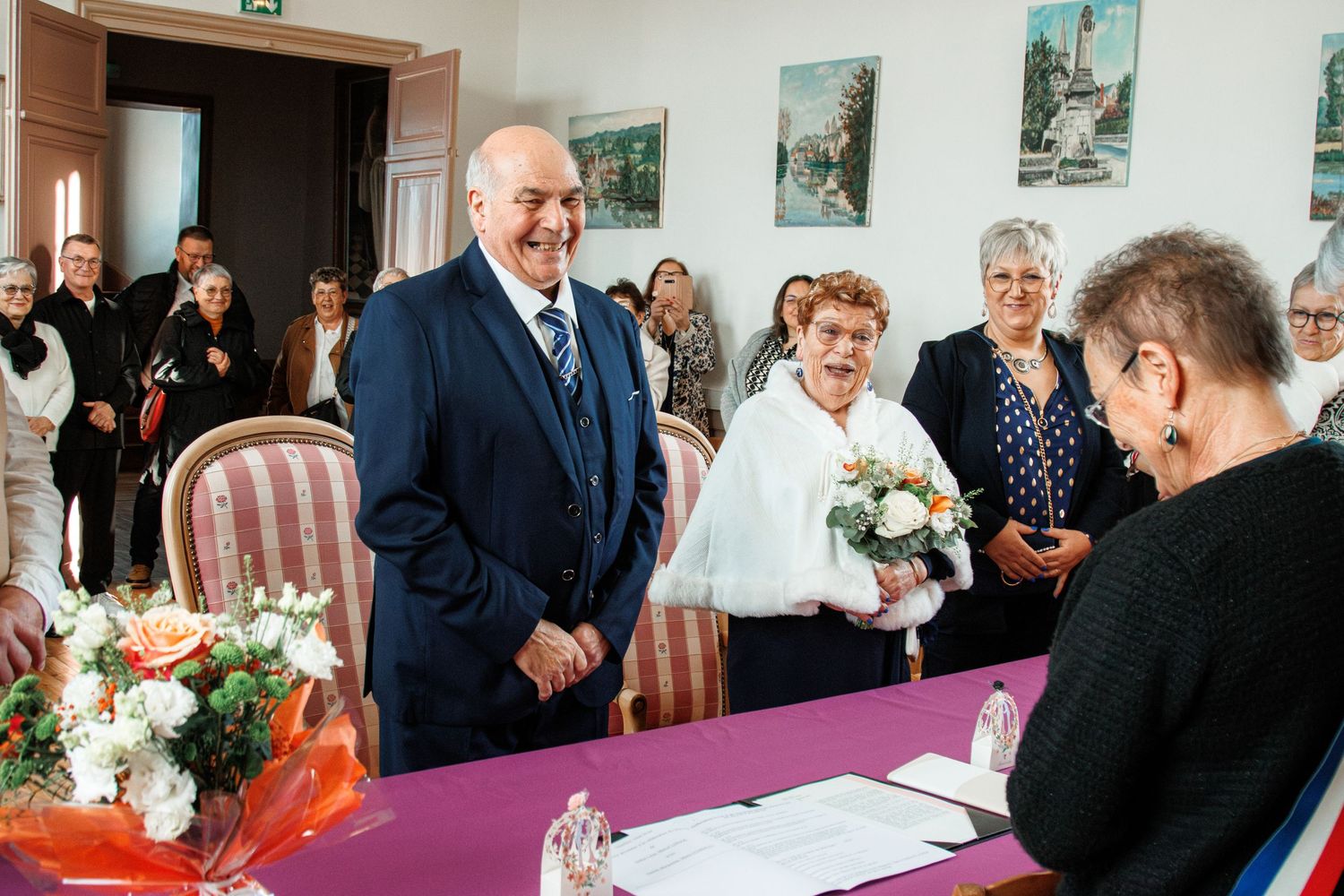 Elderly couple smiling during a civil wedding ceremony indoors, with guests and a registrar at a decorated table.