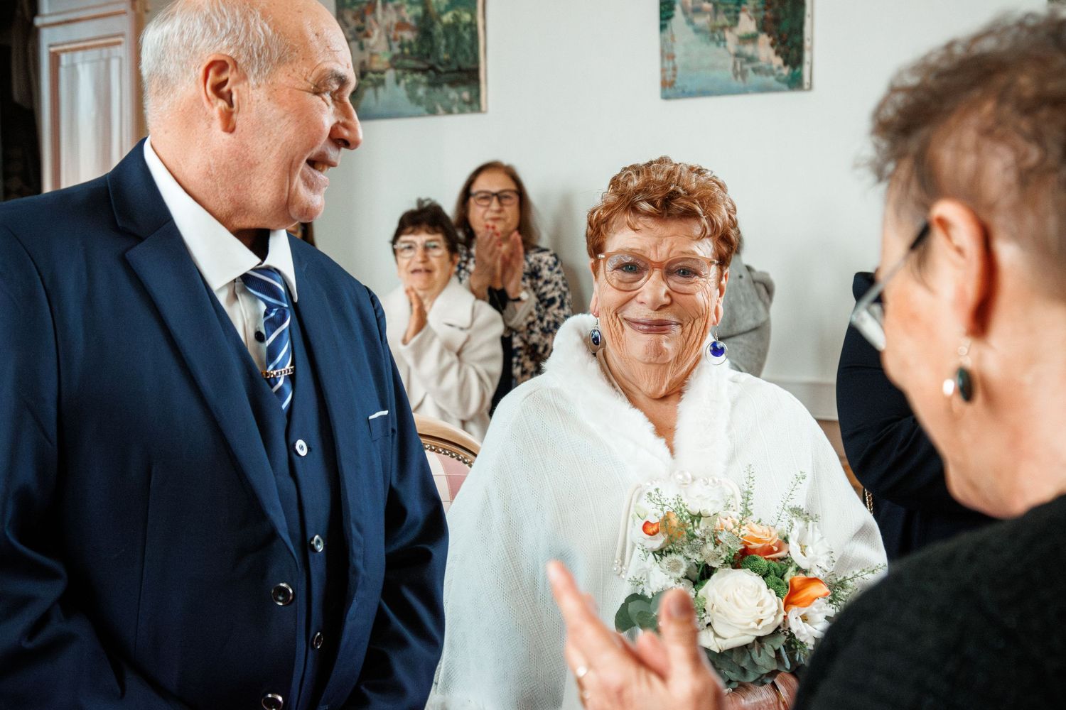 Elderly couple celebrating wedding ceremony, bride holding bouquet, guests applauding indoors.
