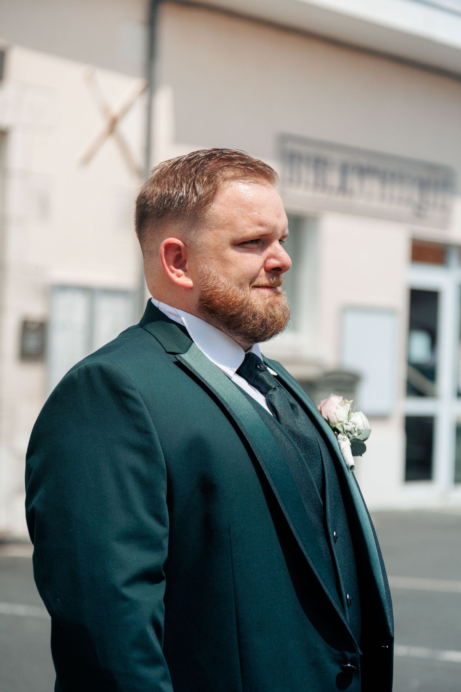 Bearded groom in dark green three-piece suit with floral boutonniere poses outdoors on wedding day.