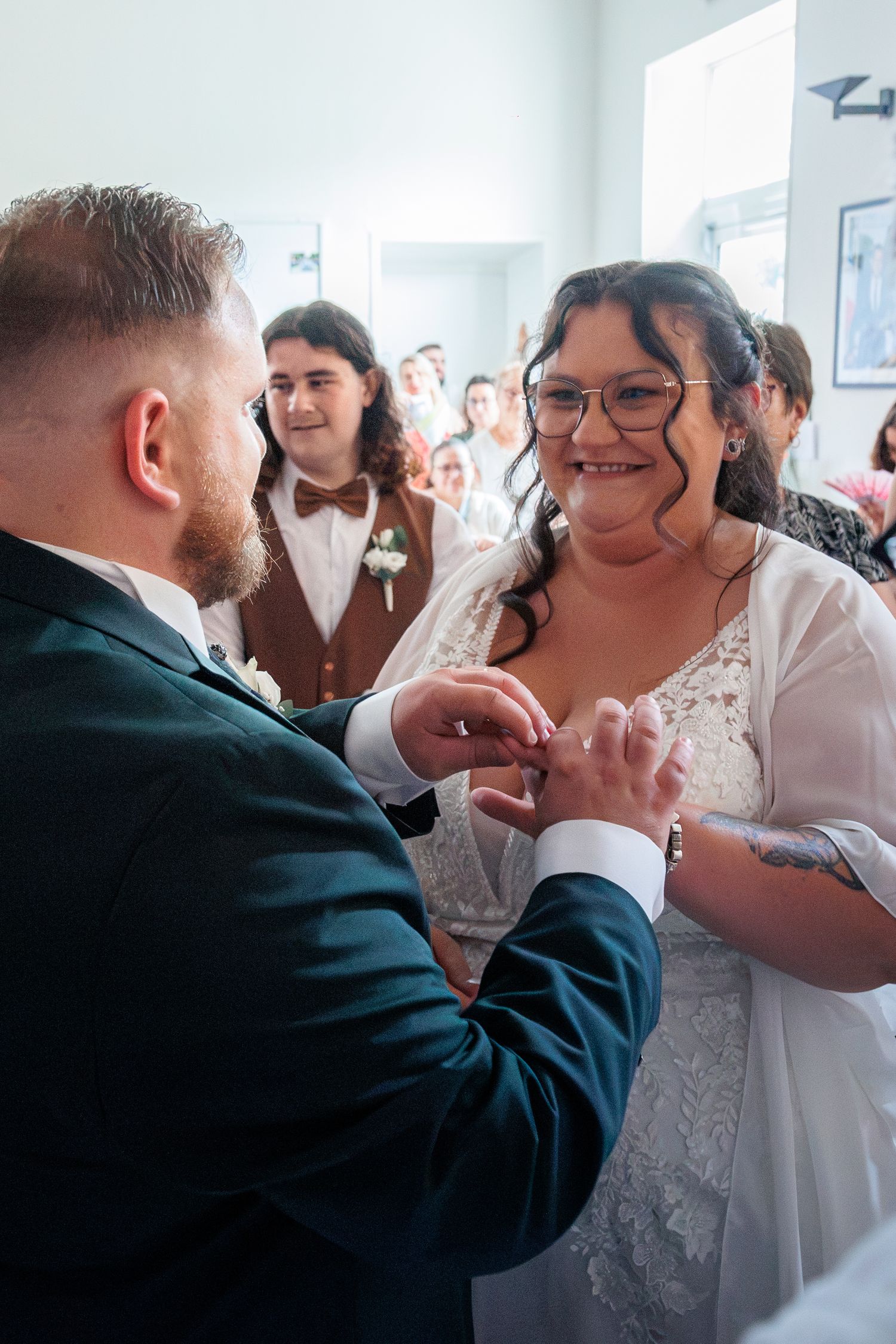 Smiling bride with glasses receives wedding ring from groom during indoor ceremony.