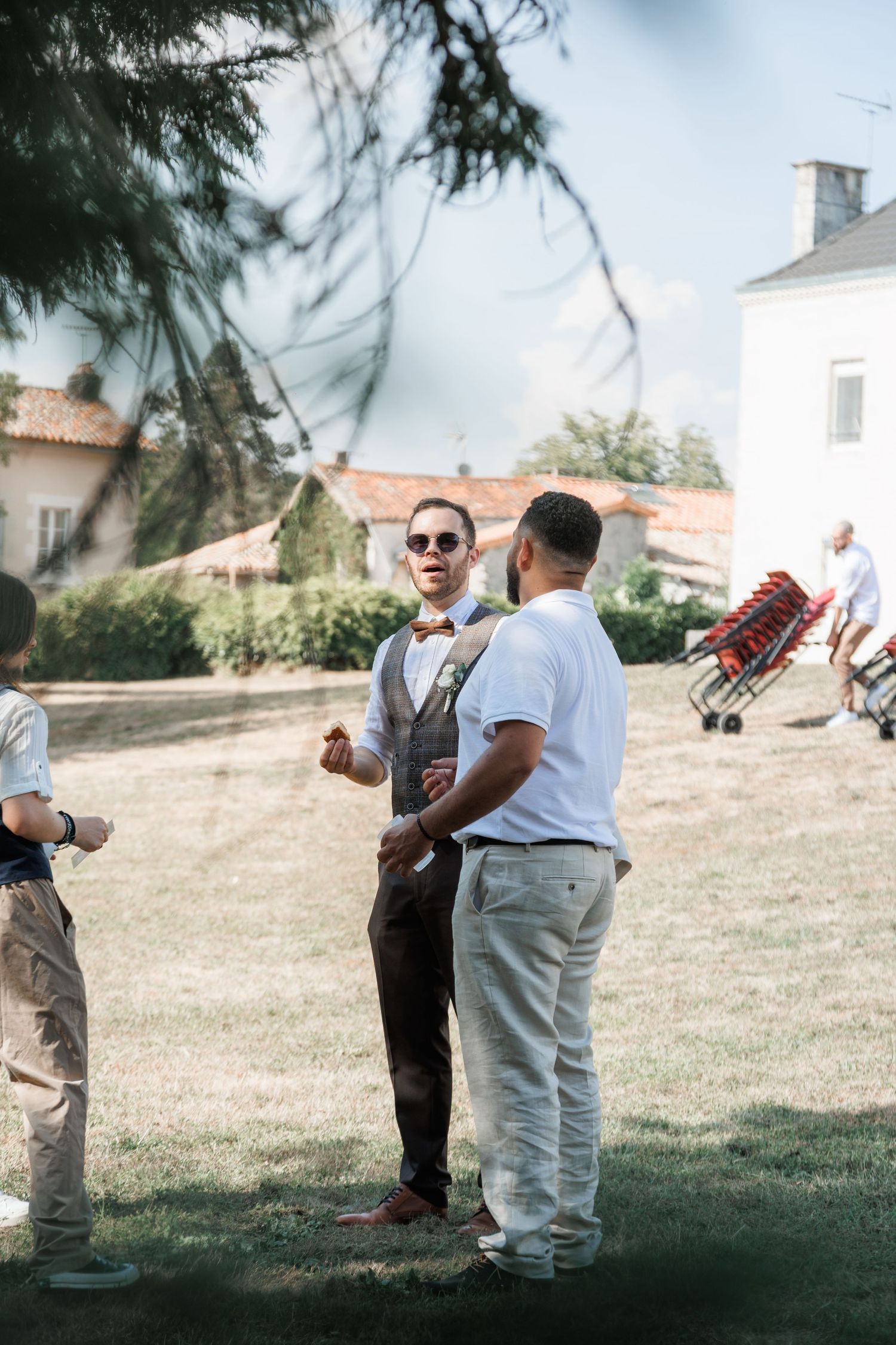 Wedding guest in vest and bow tie wearing sunglasses converses with another guest on garden lawn.