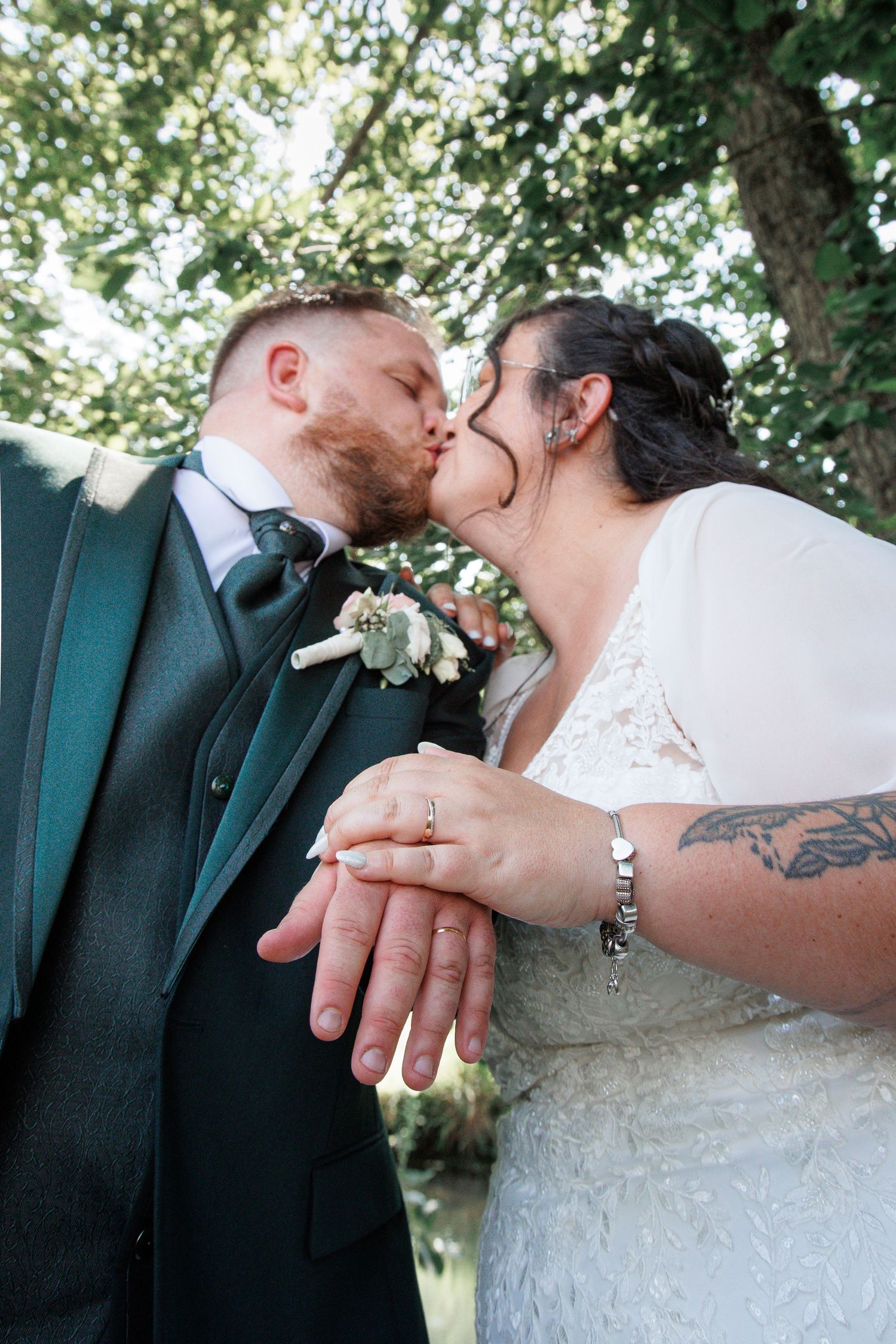 Newlyweds kissing while displaying their wedding rings outdoors under a green tree after their ceremony.