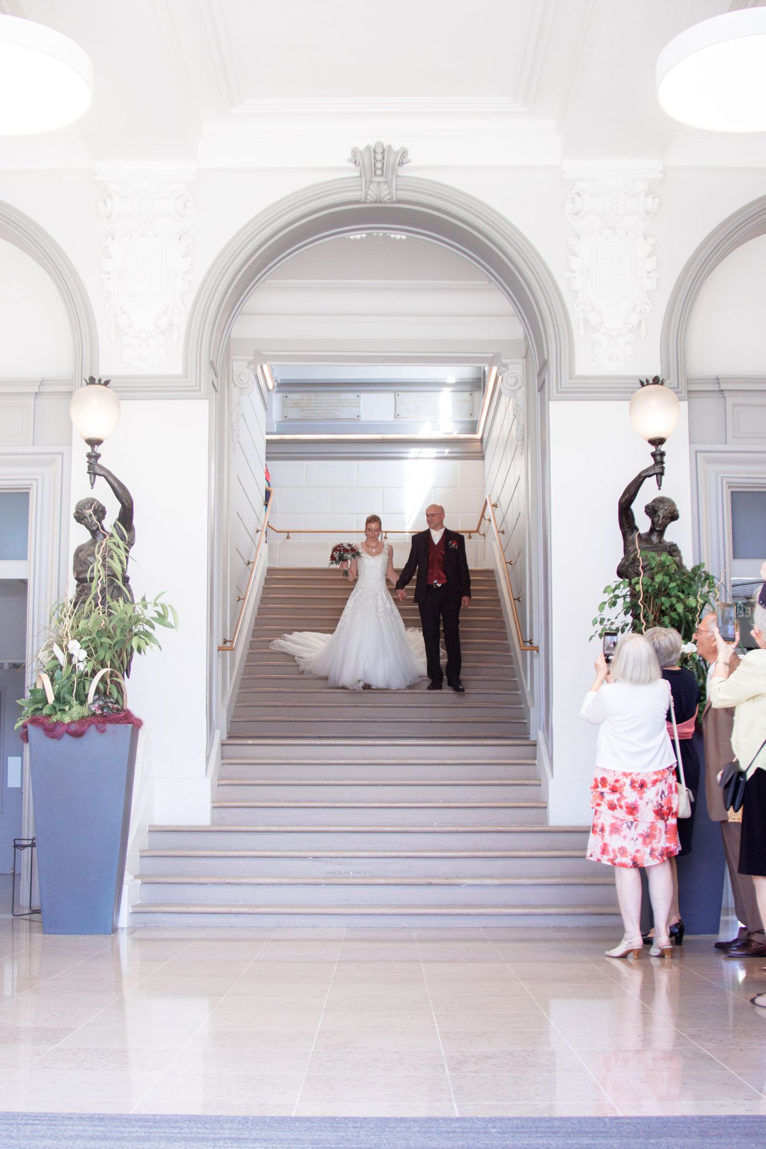 Bride and groom descend ornate staircase through arched entrance as guests photograph wedding ceremony exit.