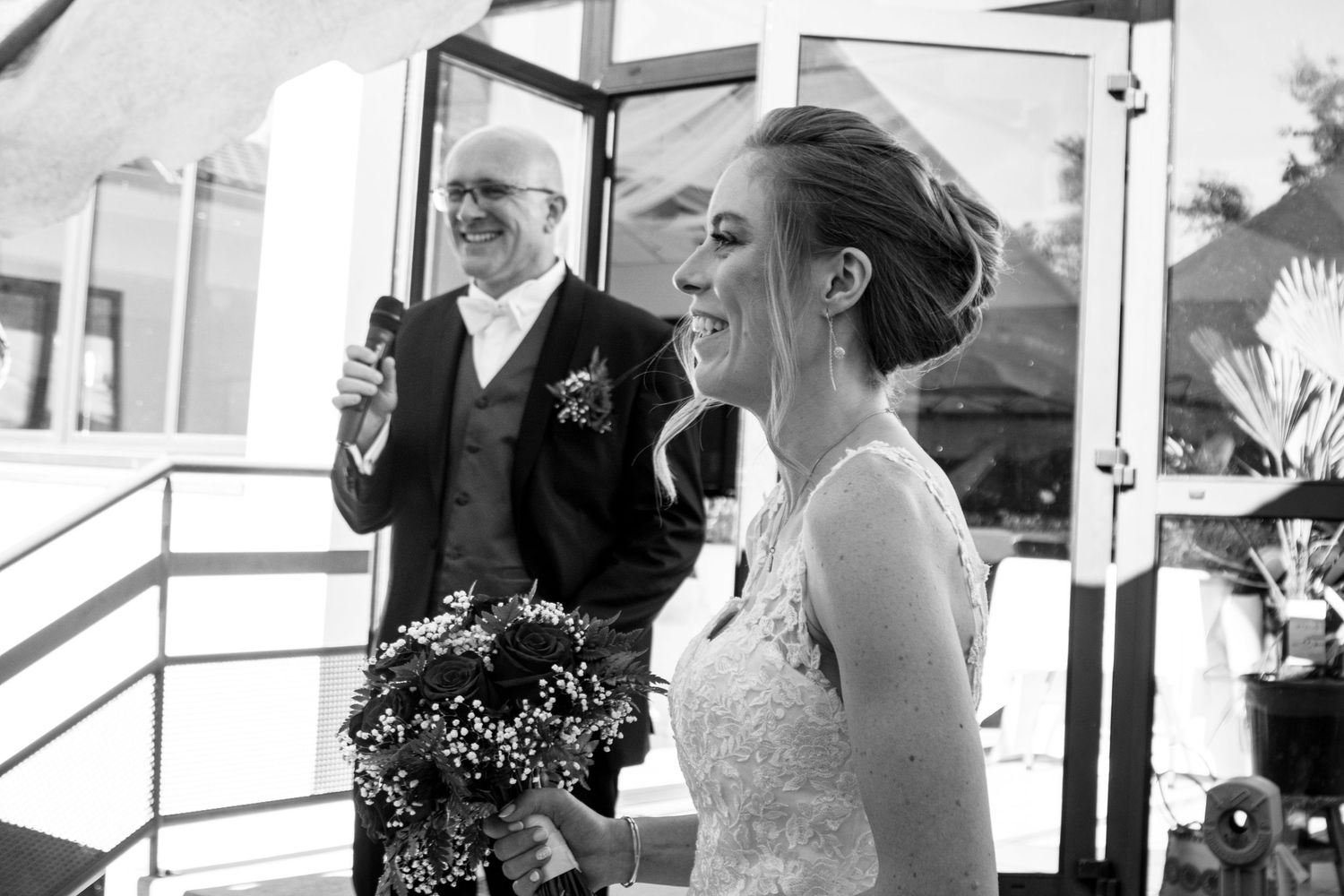 A black and white photo of a smiling bride holding a bouquet with the groom laughing behind her.