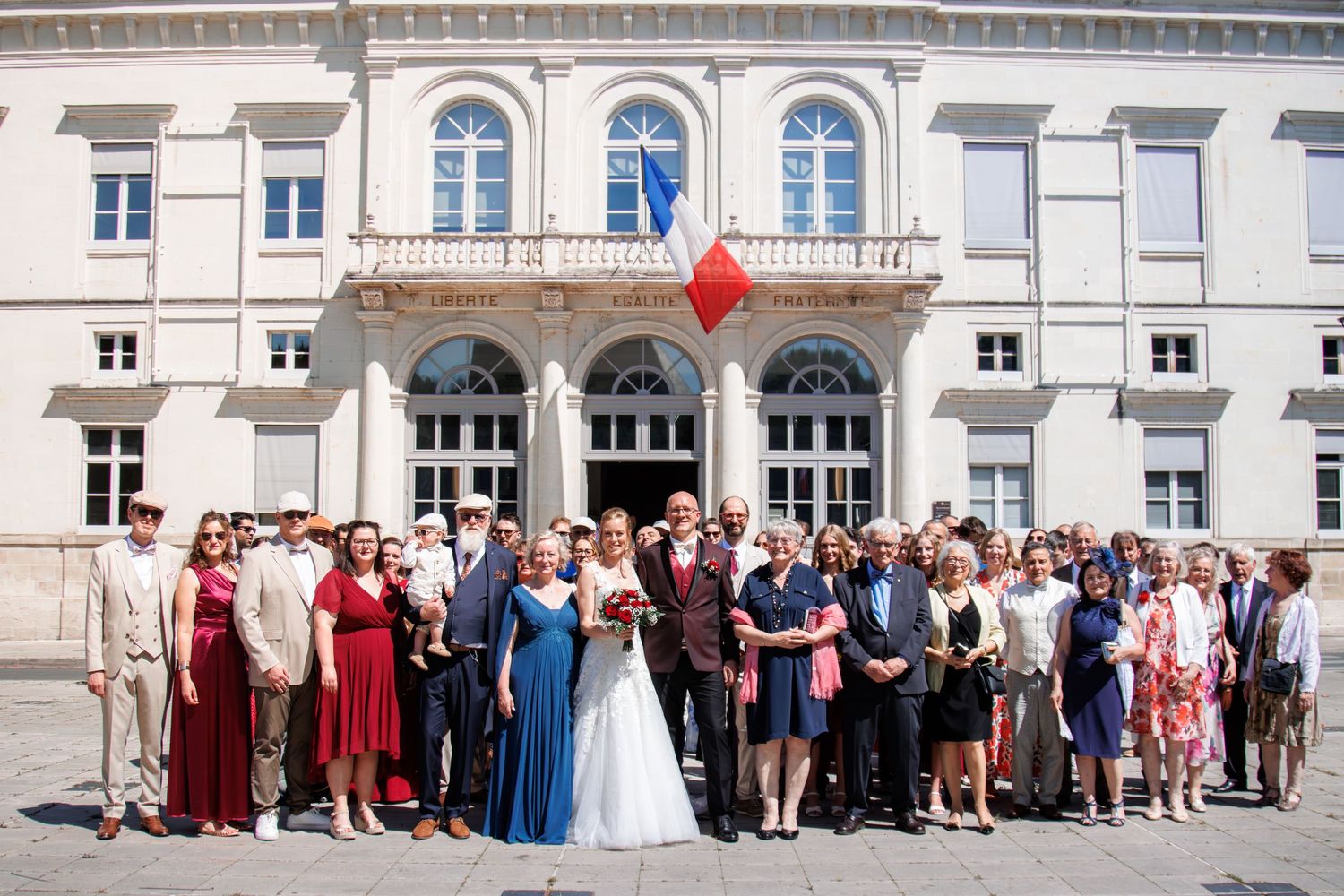 Wedding party group photo with bride and groom posing before grand white building with blue flag.