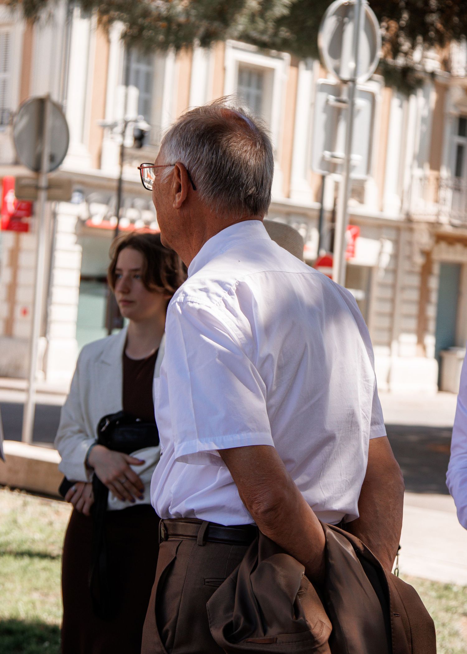 Elderly man in white shirt and glasses standing outdoors in a sunny European city square.