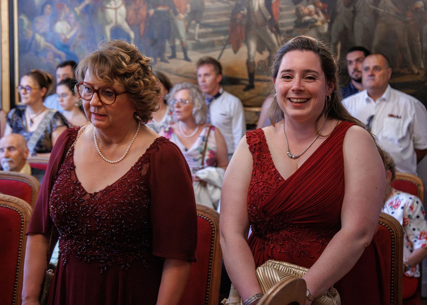 Two women in matching burgundy dresses smile during a wedding ceremony indoors.