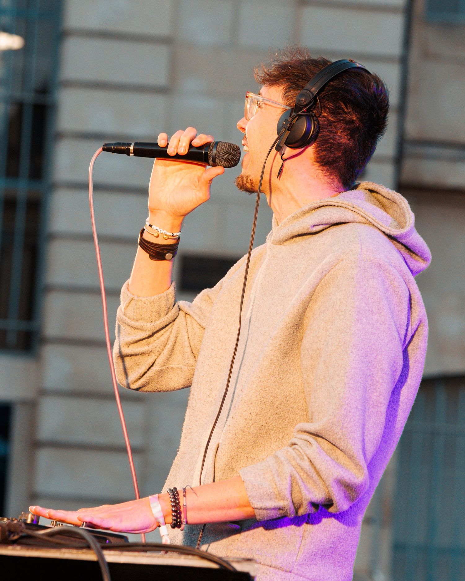 Musician in gray hoodie performs with microphone and headphones under purple stage lighting.