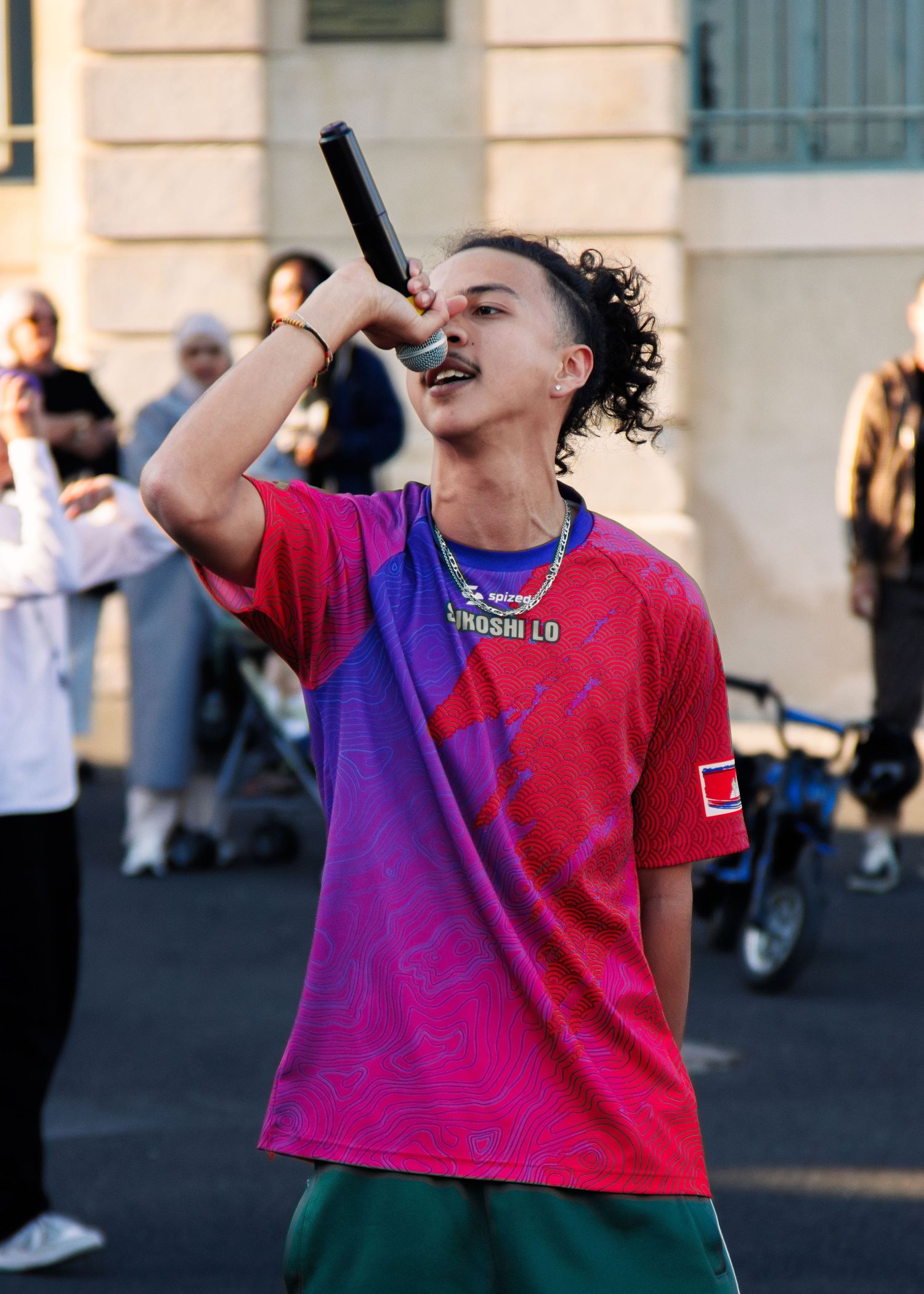 Performer in red and purple shirt holds microphone during outdoor performance.