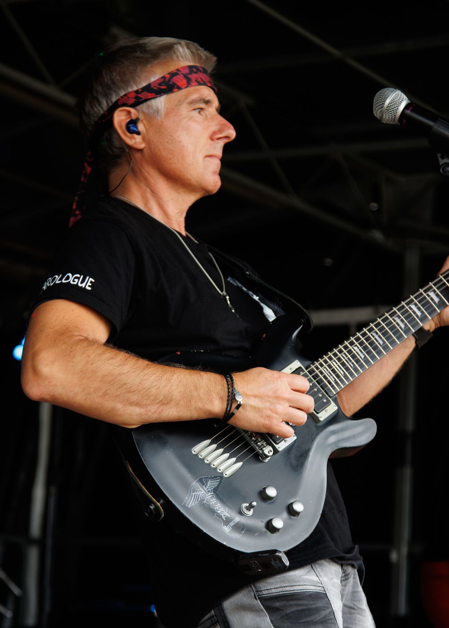 Musician performs with a gray electric guitar wearing red headband and in-ear monitors on stage.