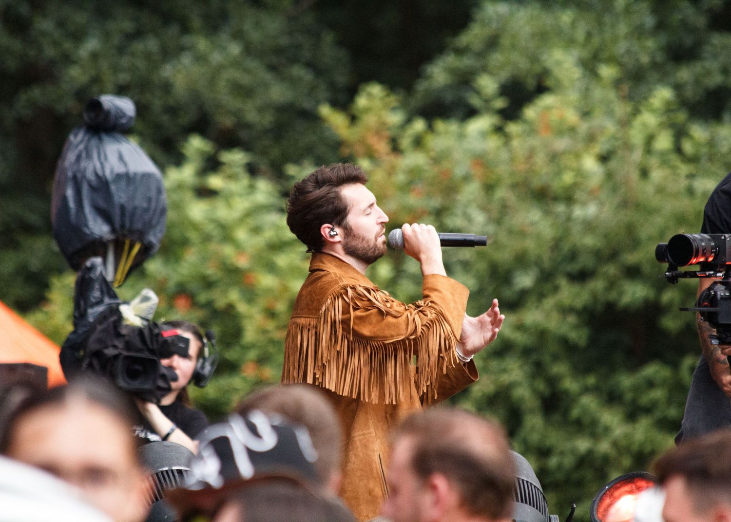 A performer in a brown fringed leather jacket sings on stage while surrounded by cameras and audience members.