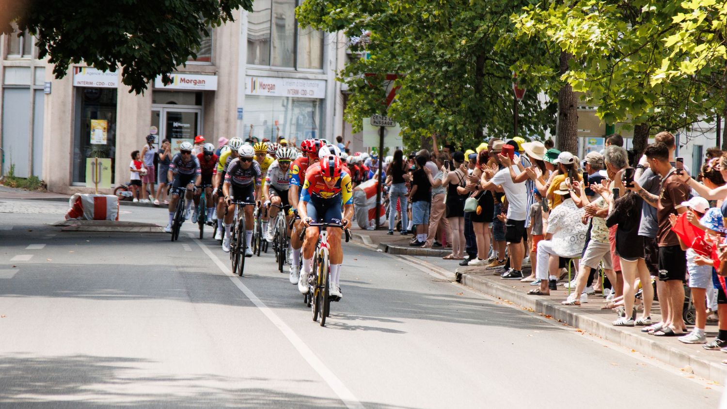 Crowds of spectators line both sides of a tree-shaded street during an outdoor event.