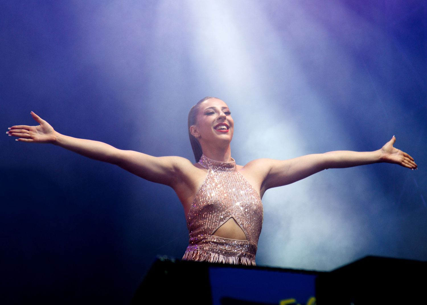 A performer in a sparkly pink dress stands with arms outstretched under dramatic blue stage lighting.