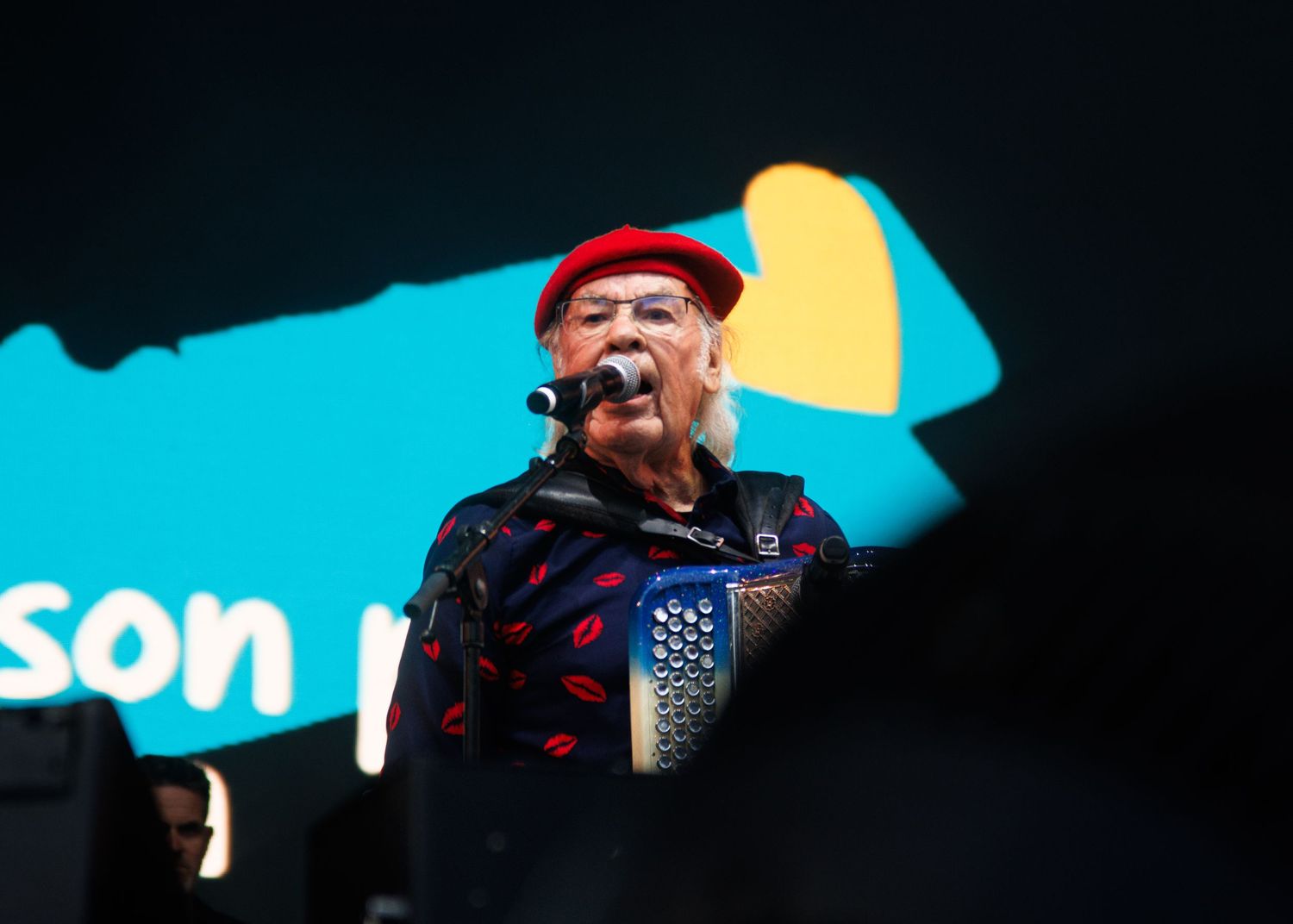 A performer in a red beret sings passionately on stage against a vibrant turquoise backdrop with yellow accents.