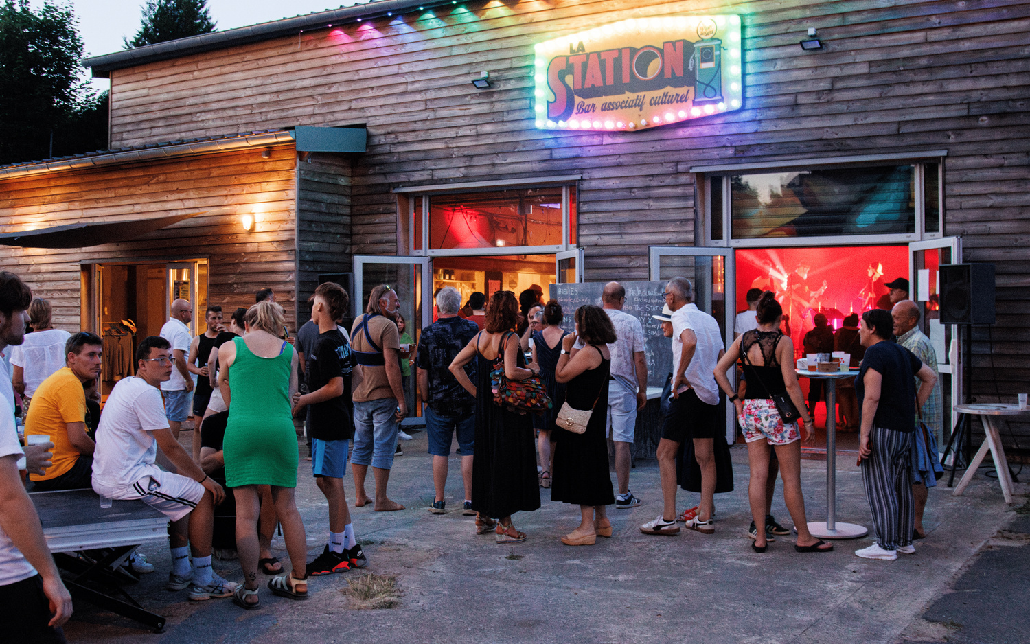 People line up outside a wooden building with a neon rainbow sign reading 'Santos' in the evening.