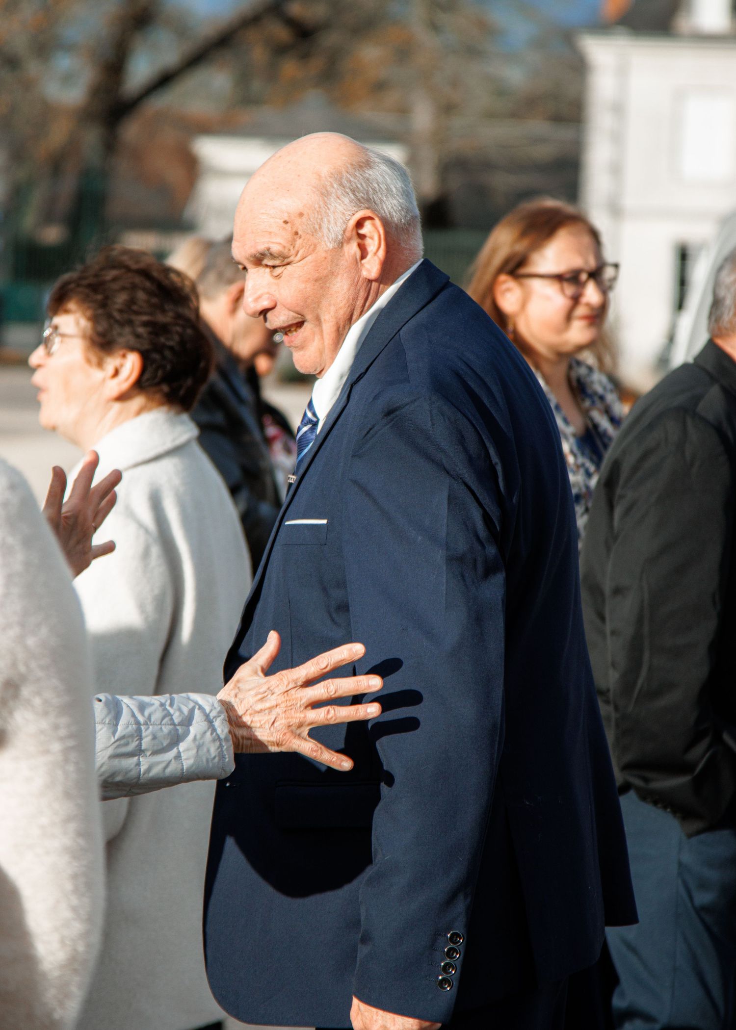 Elderly bald man in navy suit gesturing outdoors among a crowd of people on a sunny day.
