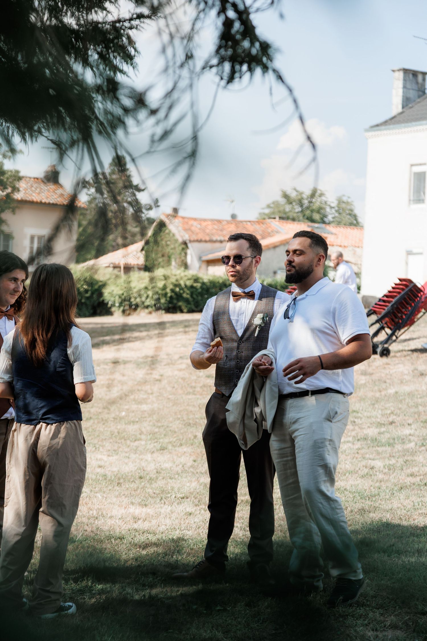 Groom in tweed vest and bow tie with sunglasses chats with guests on a sunny lawn at an outdoor wedding.