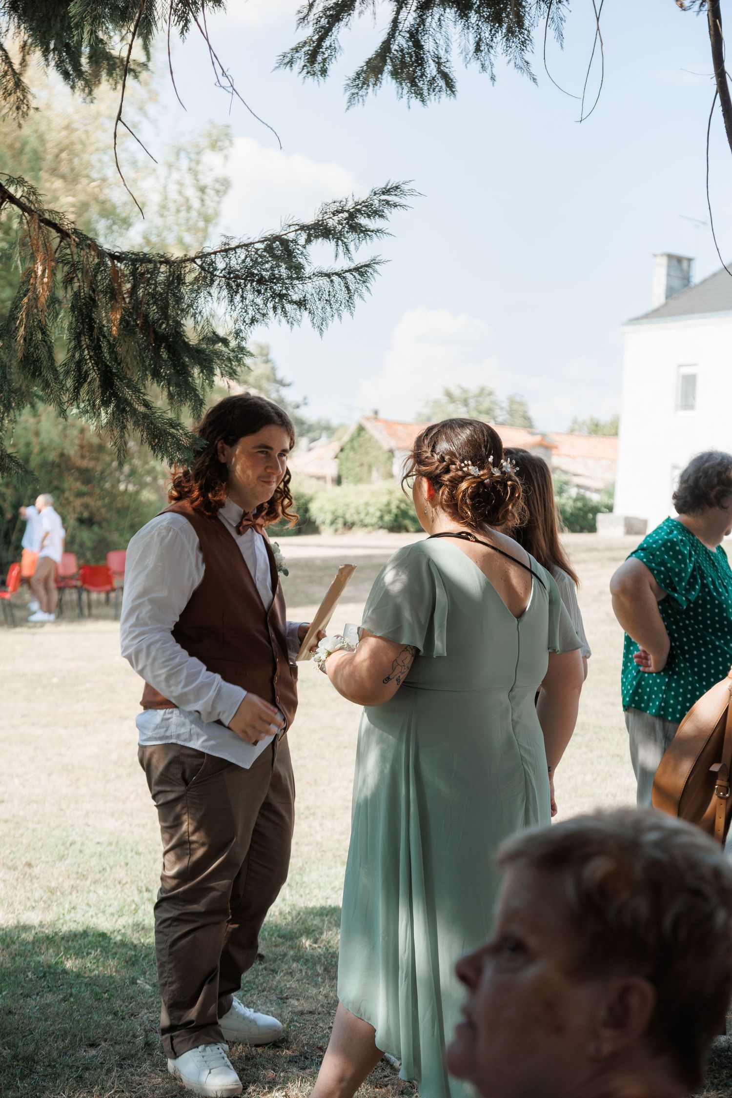 Guests mingle outdoors at a summer wedding reception, with a man in brown vest and woman in sage green dress conversing.