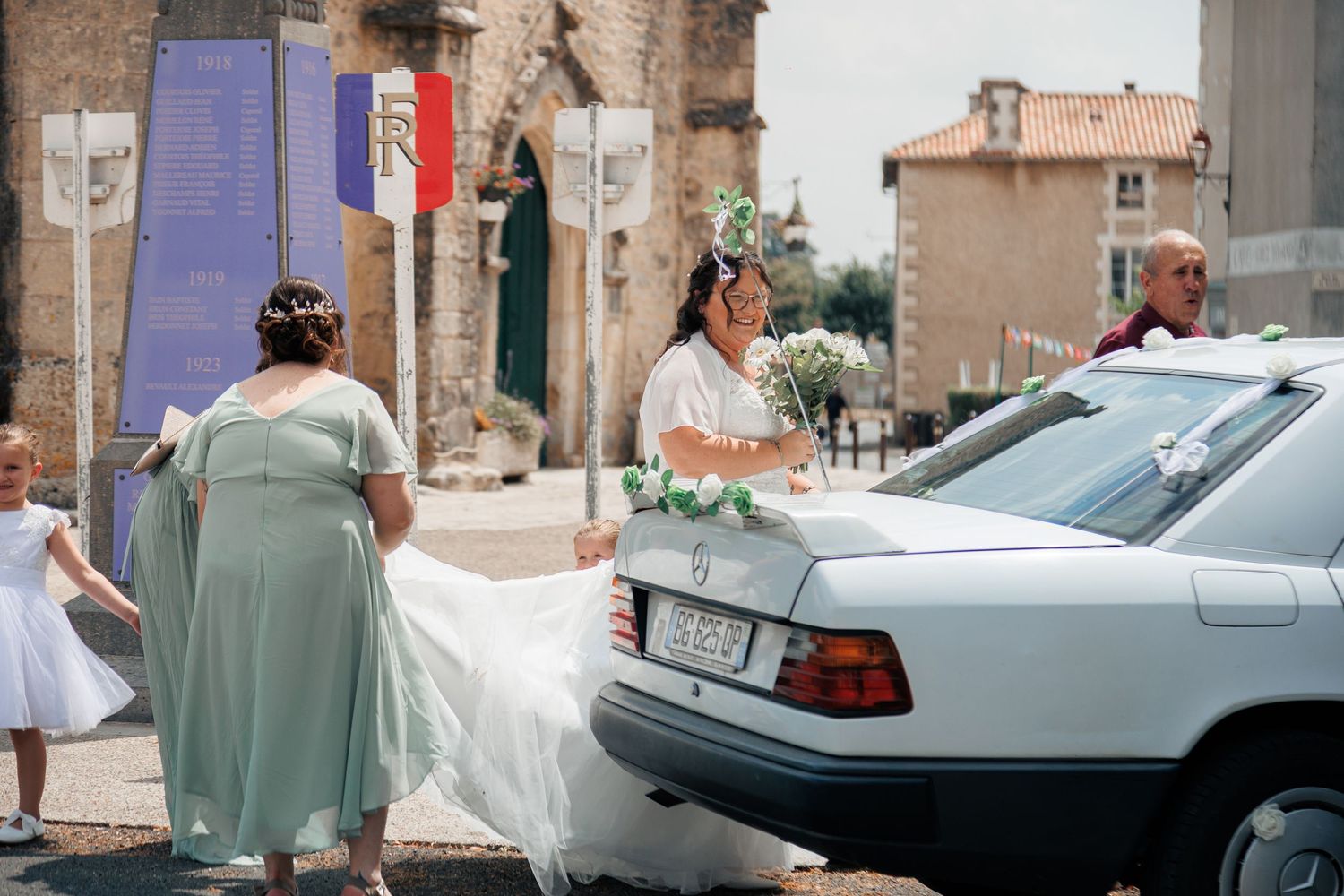 Bride in white gown arrives at wedding venue in decorated white Mercedes, assisted by bridesmaid in sage green dress.