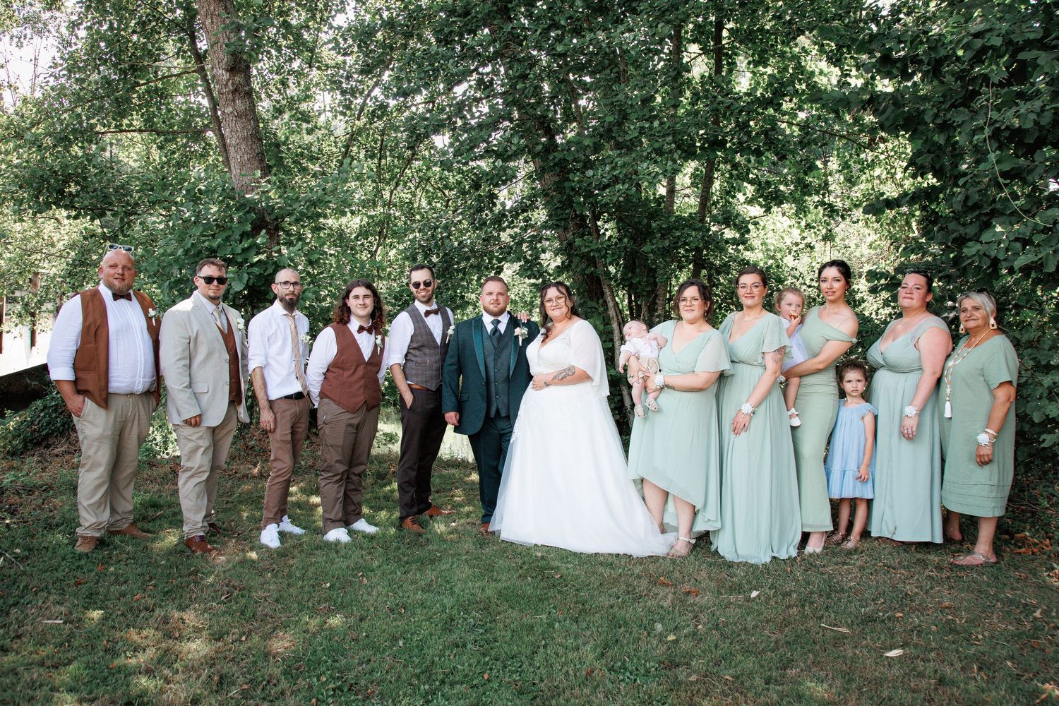 Wedding party poses outdoors under lush green trees, featuring bride, groom, groomsmen, and bridesmaids in sage green dresses.