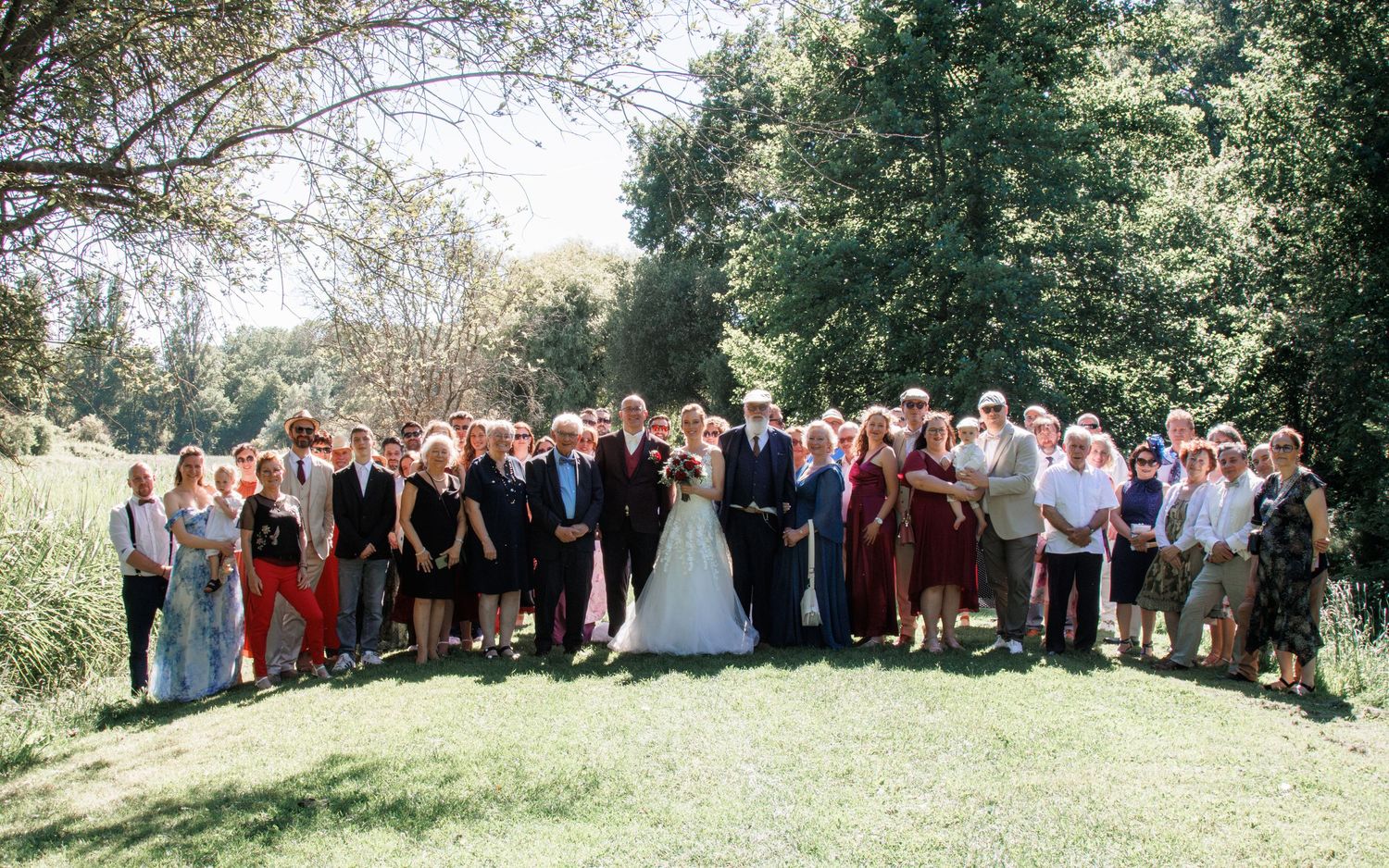Large wedding party group photo outdoors on green lawn surrounded by lush trees on a sunny day.