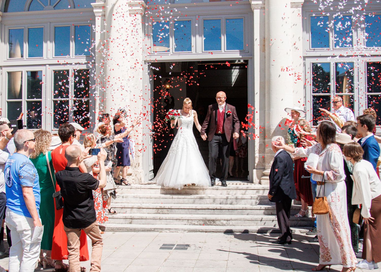 Newlyweds exit a grand building through a shower of colorful confetti, surrounded by cheering wedding guests.