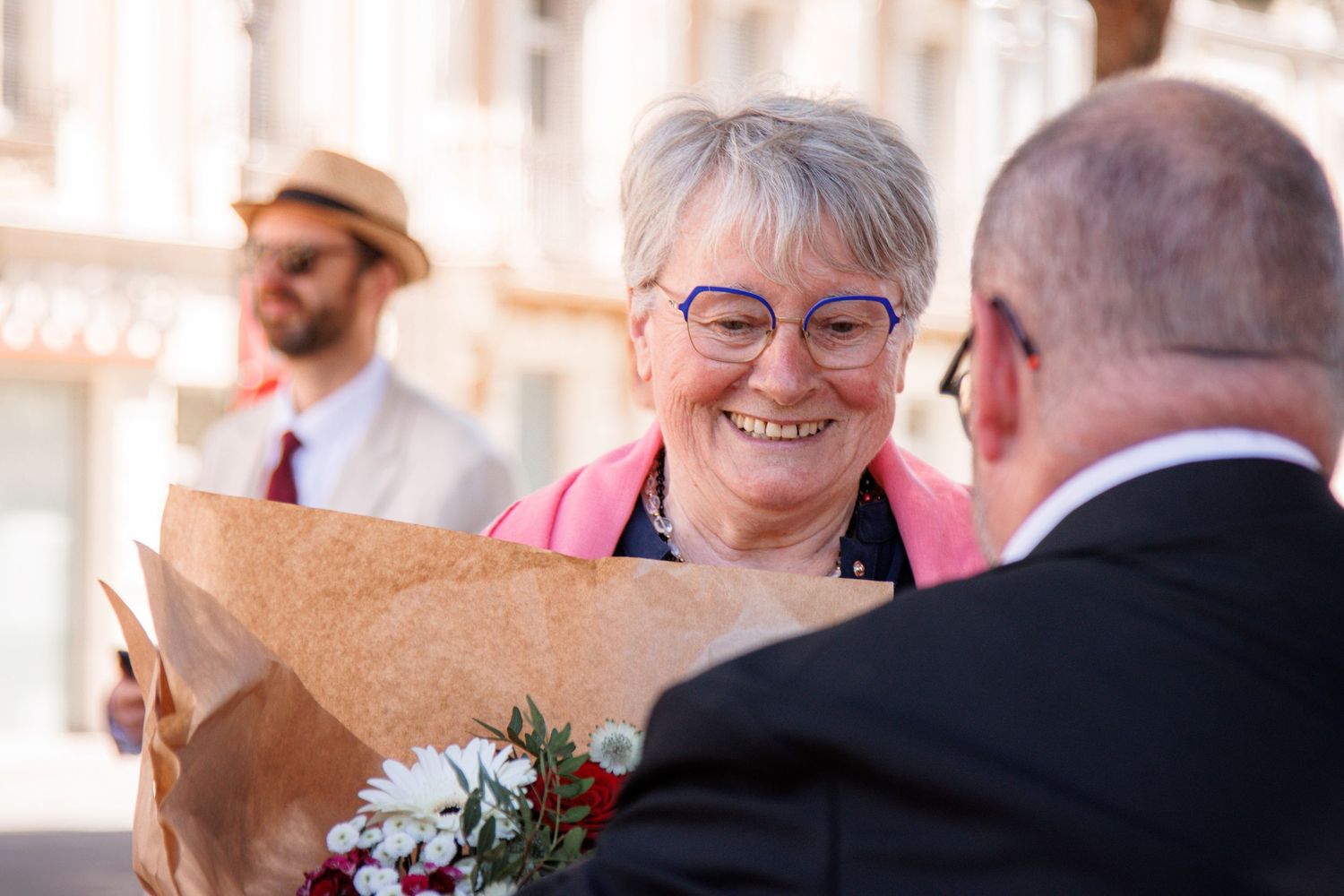 Smiling elderly woman with glasses receiving a bouquet of flowers outdoors at a sunny event.