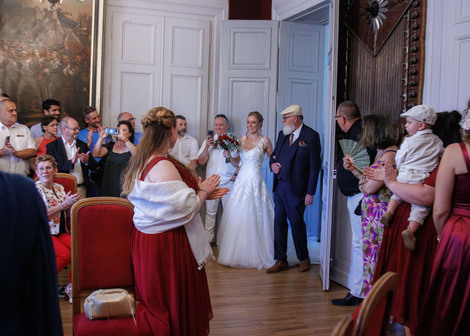 Bride in white gown enters ornate wedding hall with groom amid applauding guests.