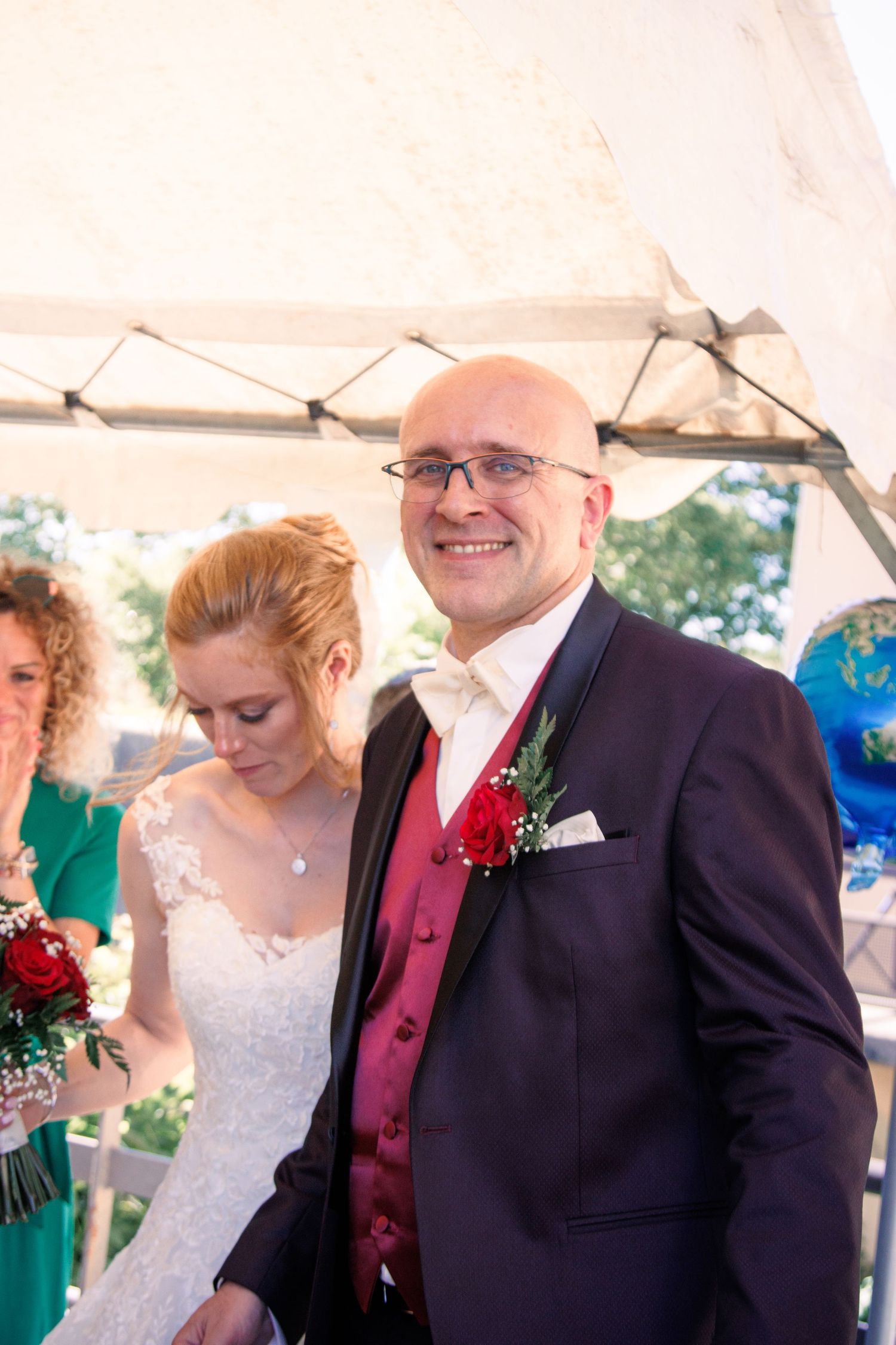 Smiling bald groom in dark suit with red boutonniere stands beside bride in white lace wedding dress.