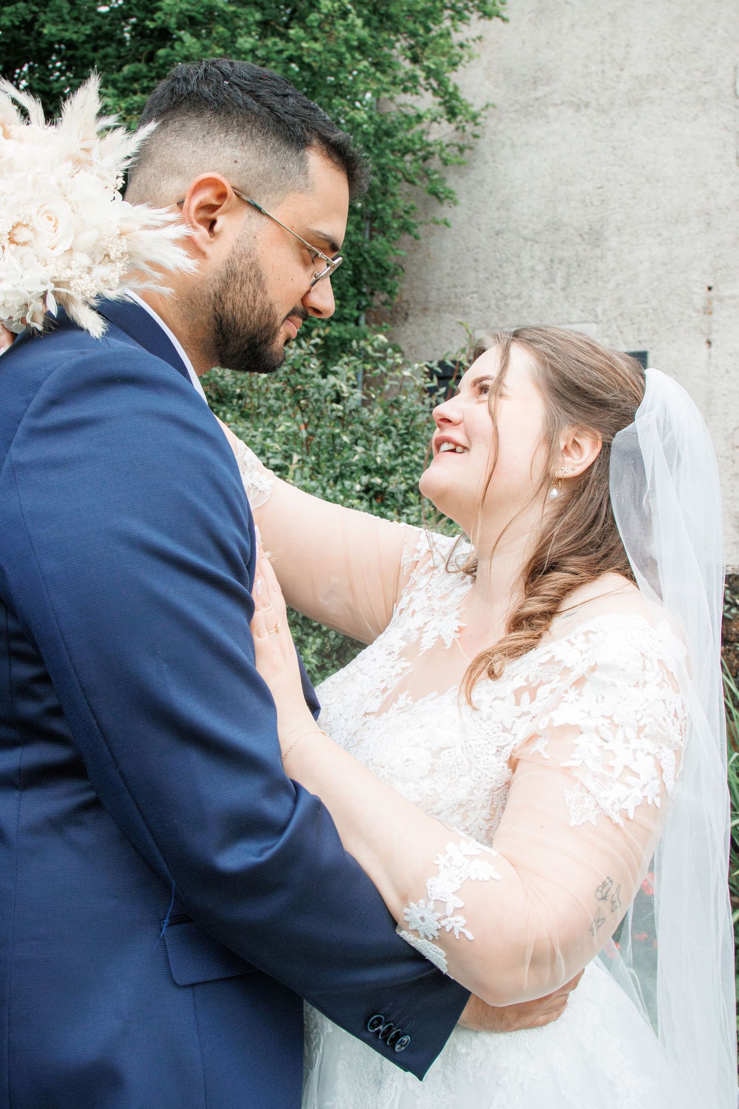 Bride and groom share a tender moment, smiling at each other, she in lace gown with veil, he in navy suit.